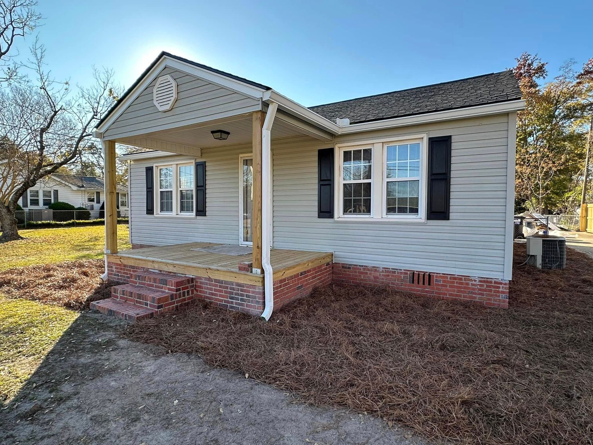 Small beige house with black shutters, white porch columns, and a brick foundation in a sunny yard