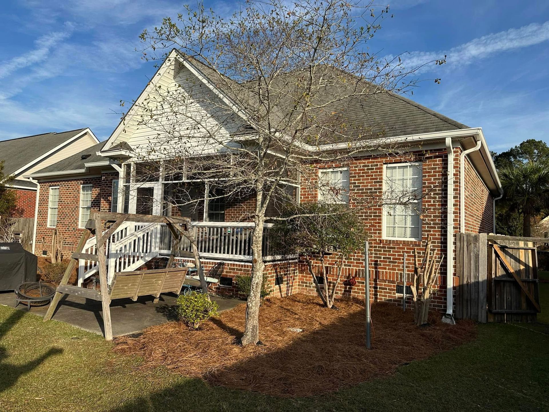Brick house with a front porch, leafless trees, and a fenced yard on a sunny day