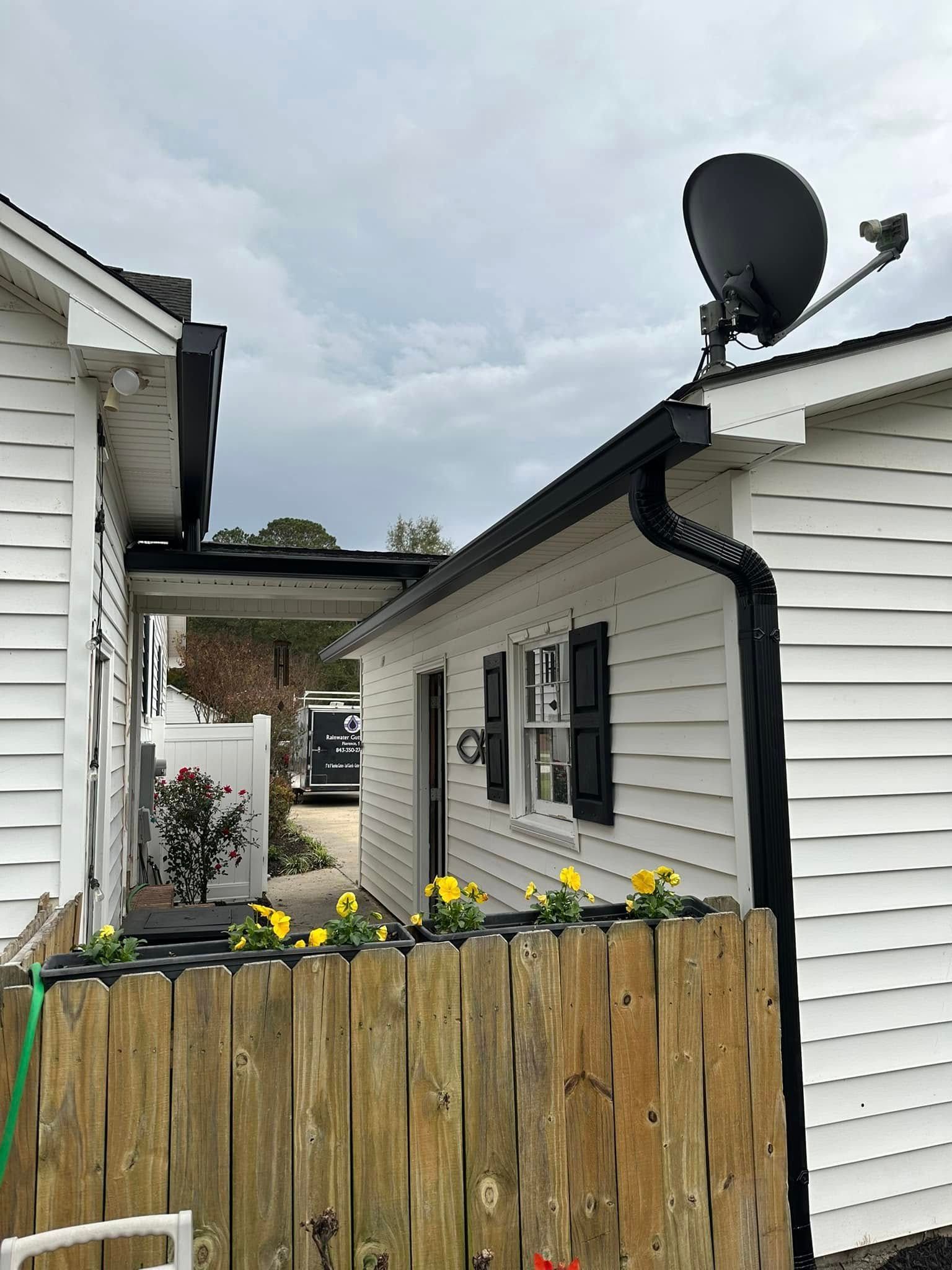 Narrow backyard between white houses with a wooden fence, yellow flowers, and a satellite dish on the roof.