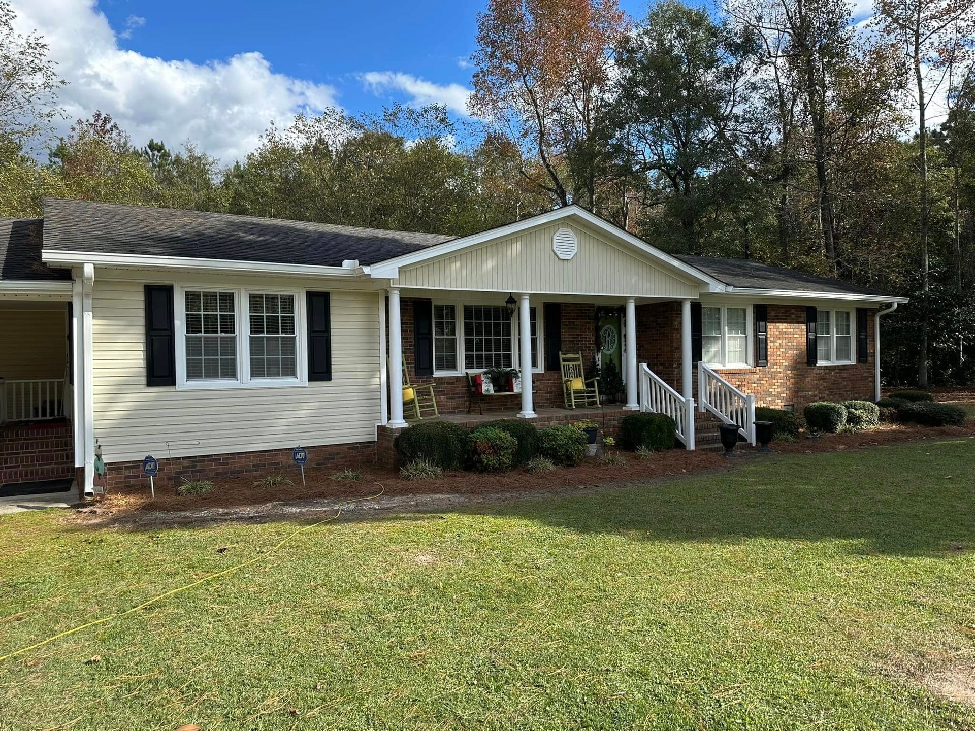 Single-story suburban house with white siding, front porch, brick accents, and a grassy yard under trees.