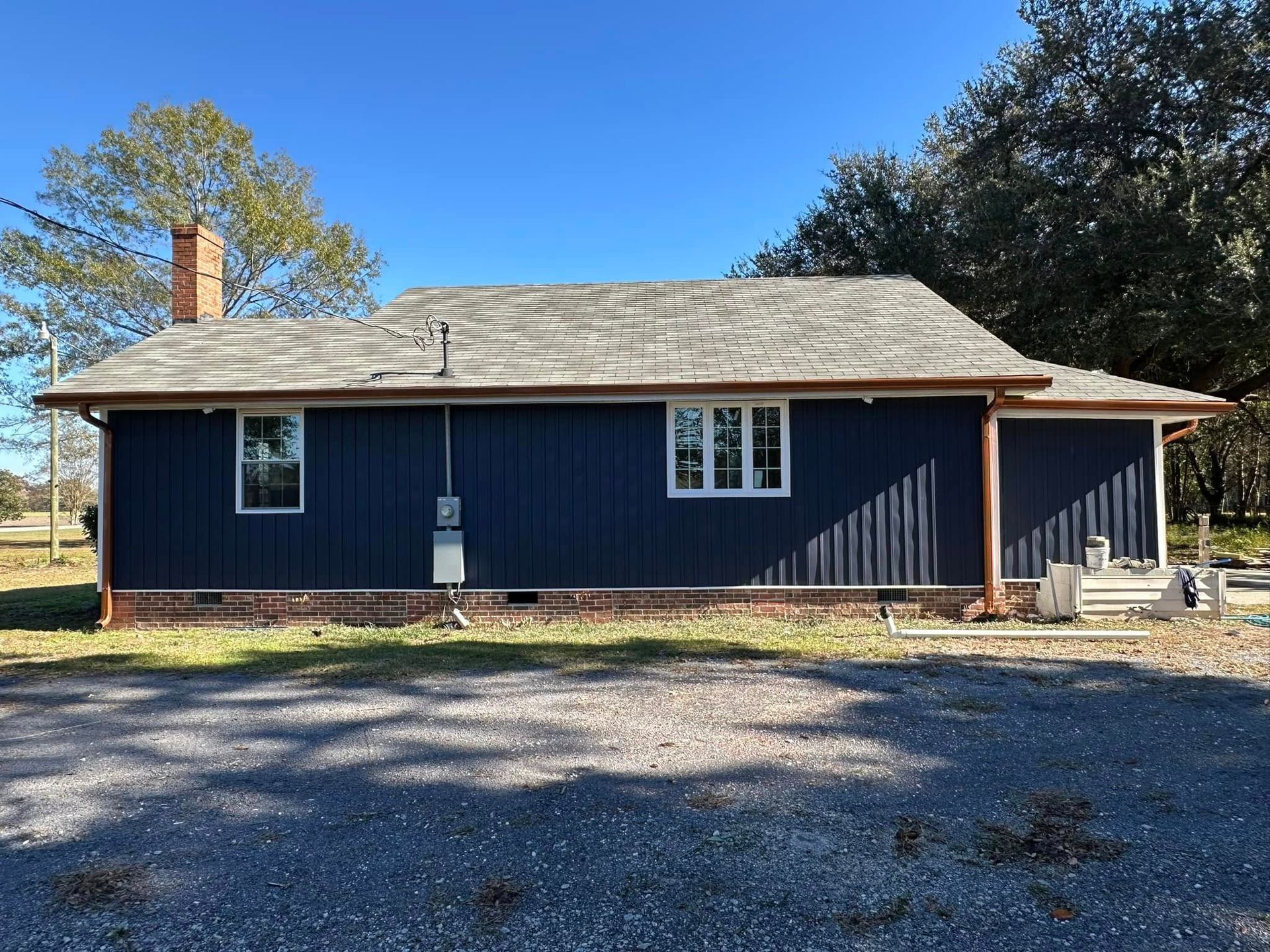 Single-story dark blue house with a tan roof, white trim, and a gravel driveway under a clear sky