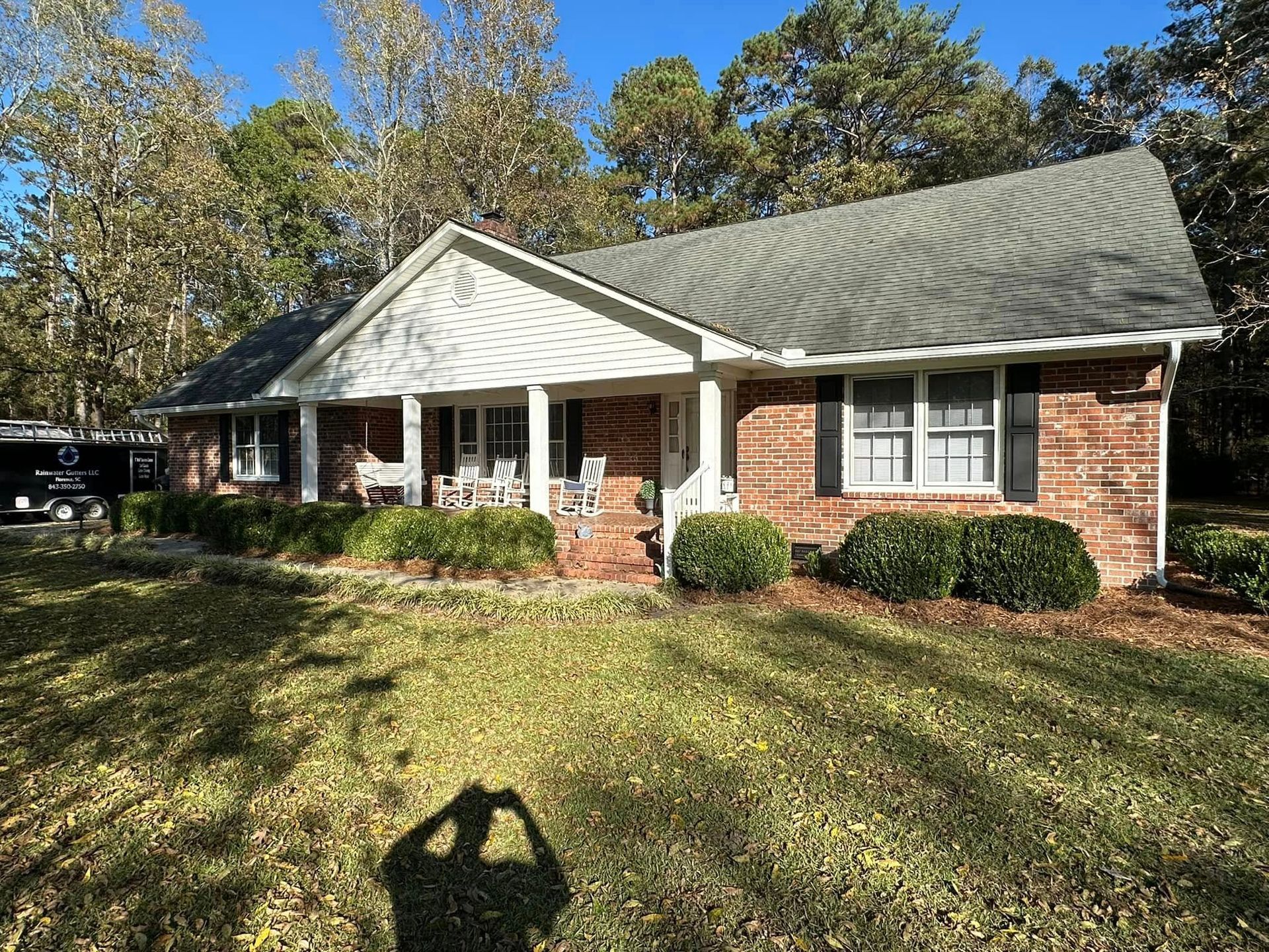 Brick ranch house with white porch and gray roof in a sunny yard with trees and bushes