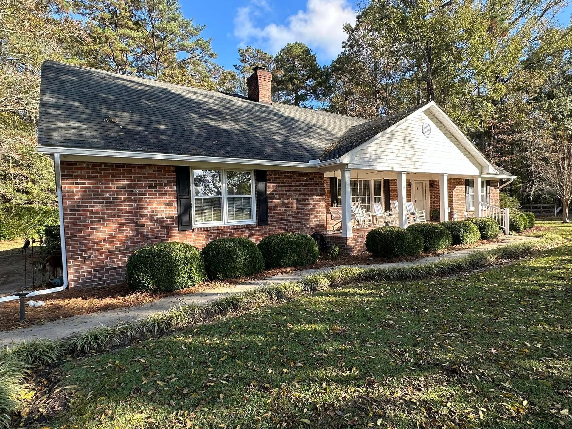 Brick house with white porch and dark roof, surrounded by trees and autumn leaves