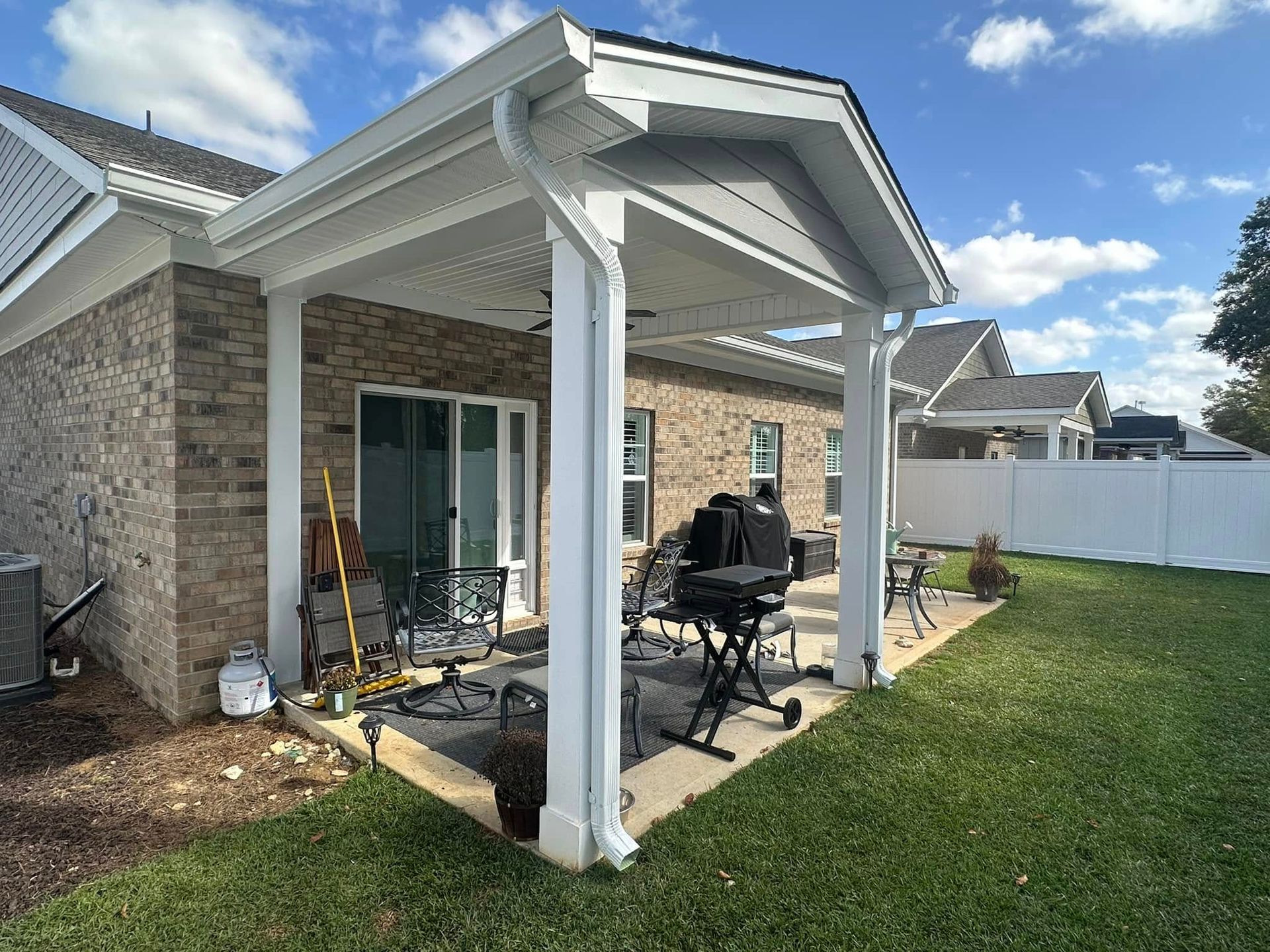 Covered patio beside a brick house, with patio furniture and a green lawn under a blue sky.