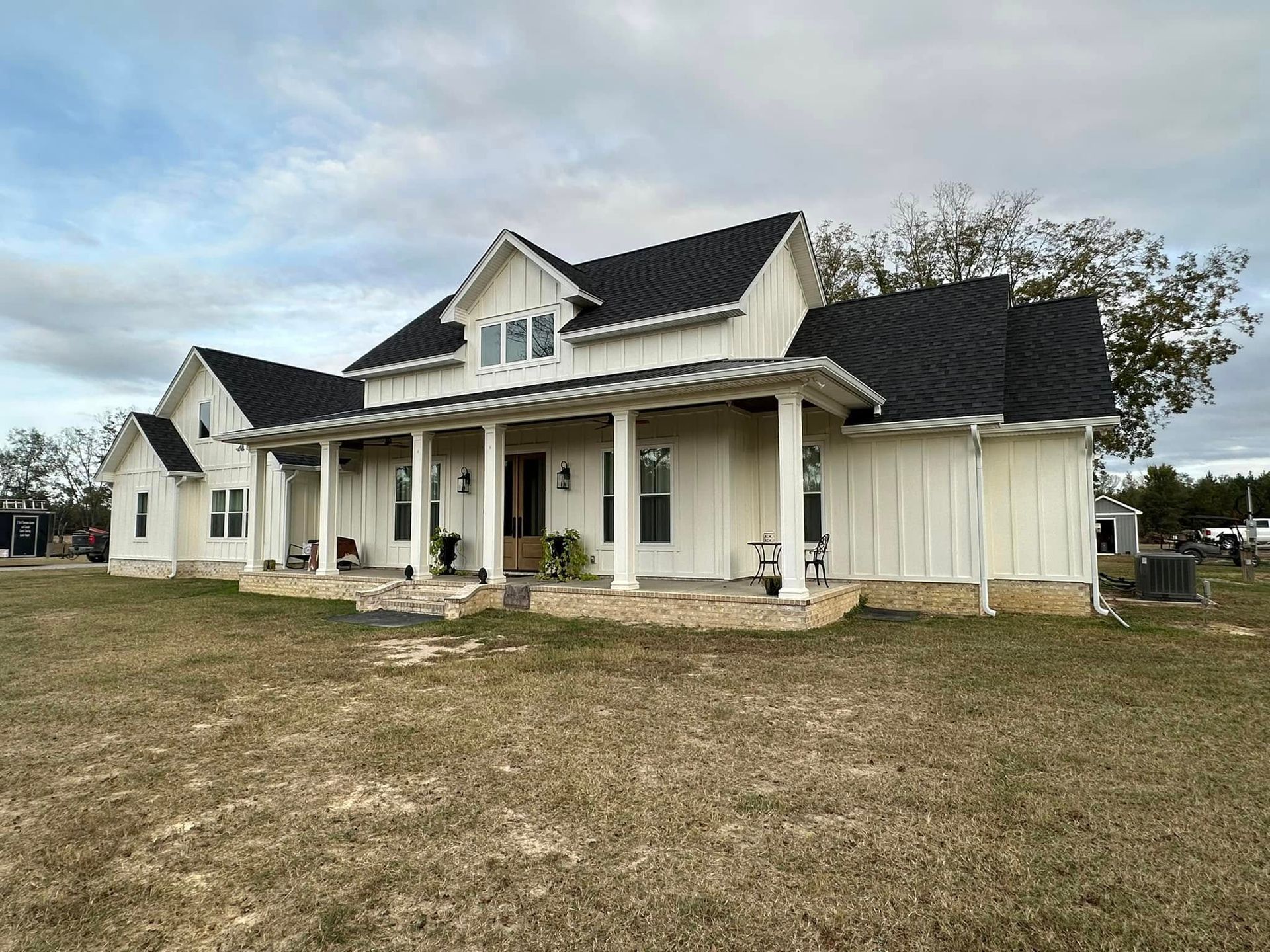 Large white farmhouse with black roof and front porch on a grassy lot under a cloudy sky