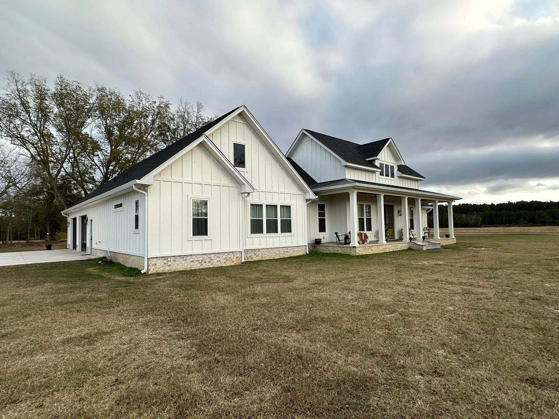 White farmhouse with porch and dark roof in a grassy field under a cloudy sky