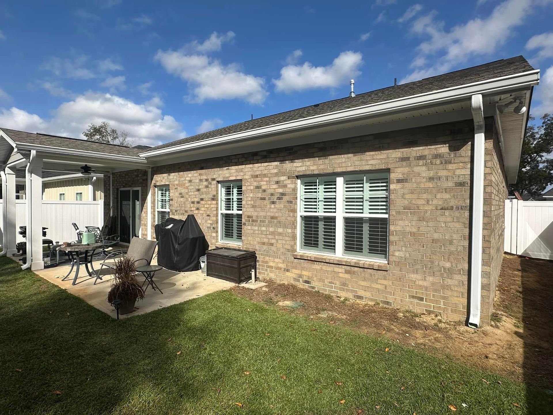 Backyard patio beside a brick house with white shutters, grill, and outdoor furniture under a blue sky