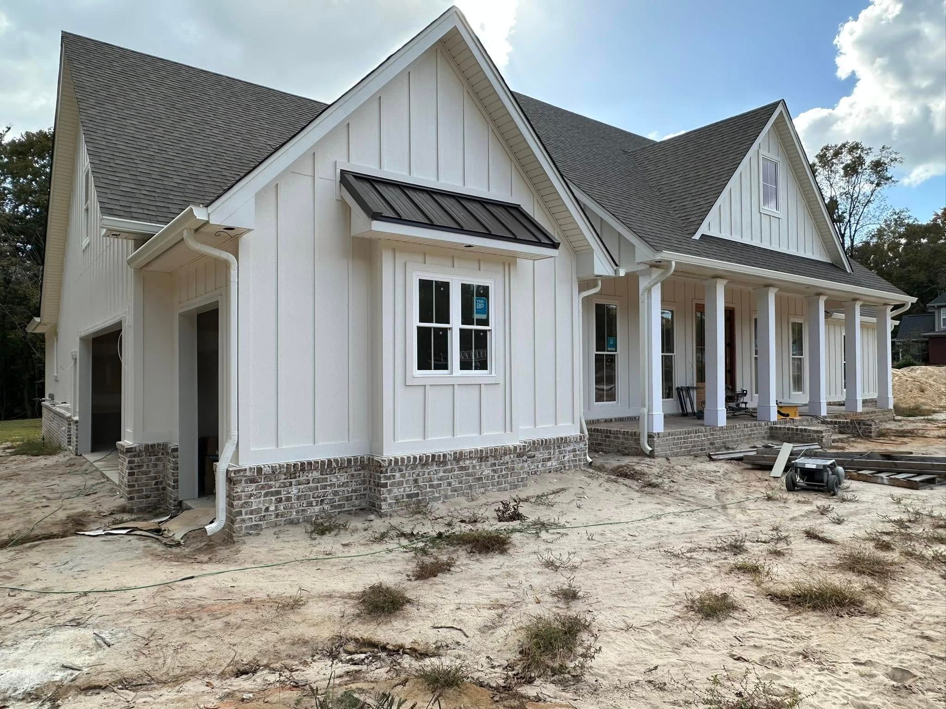 White house exterior under construction with dark gray roof and front porch, viewed from a sandy yard