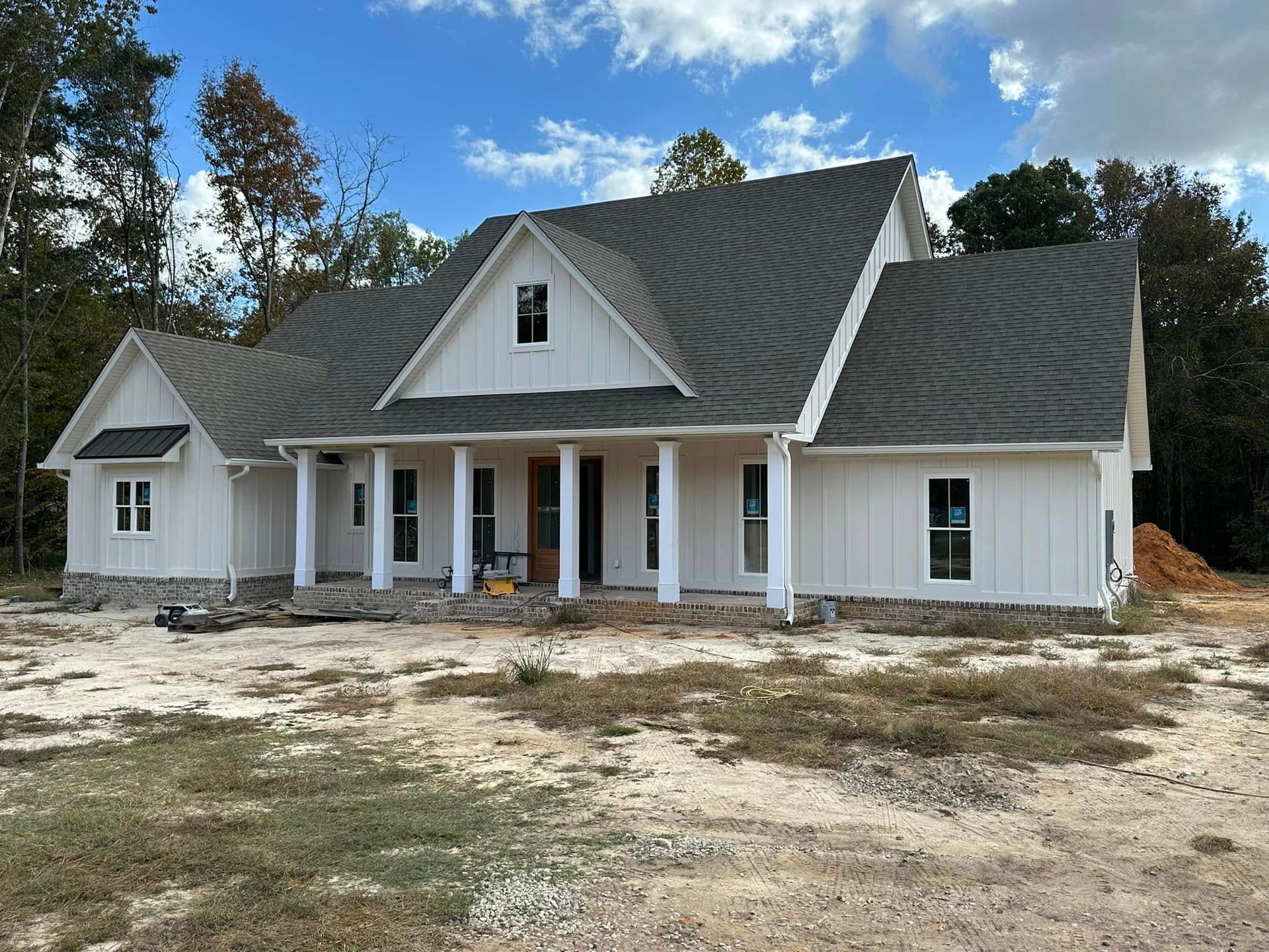White house under construction with dark gray roof, front porch, and unfinished yard under a blue sky