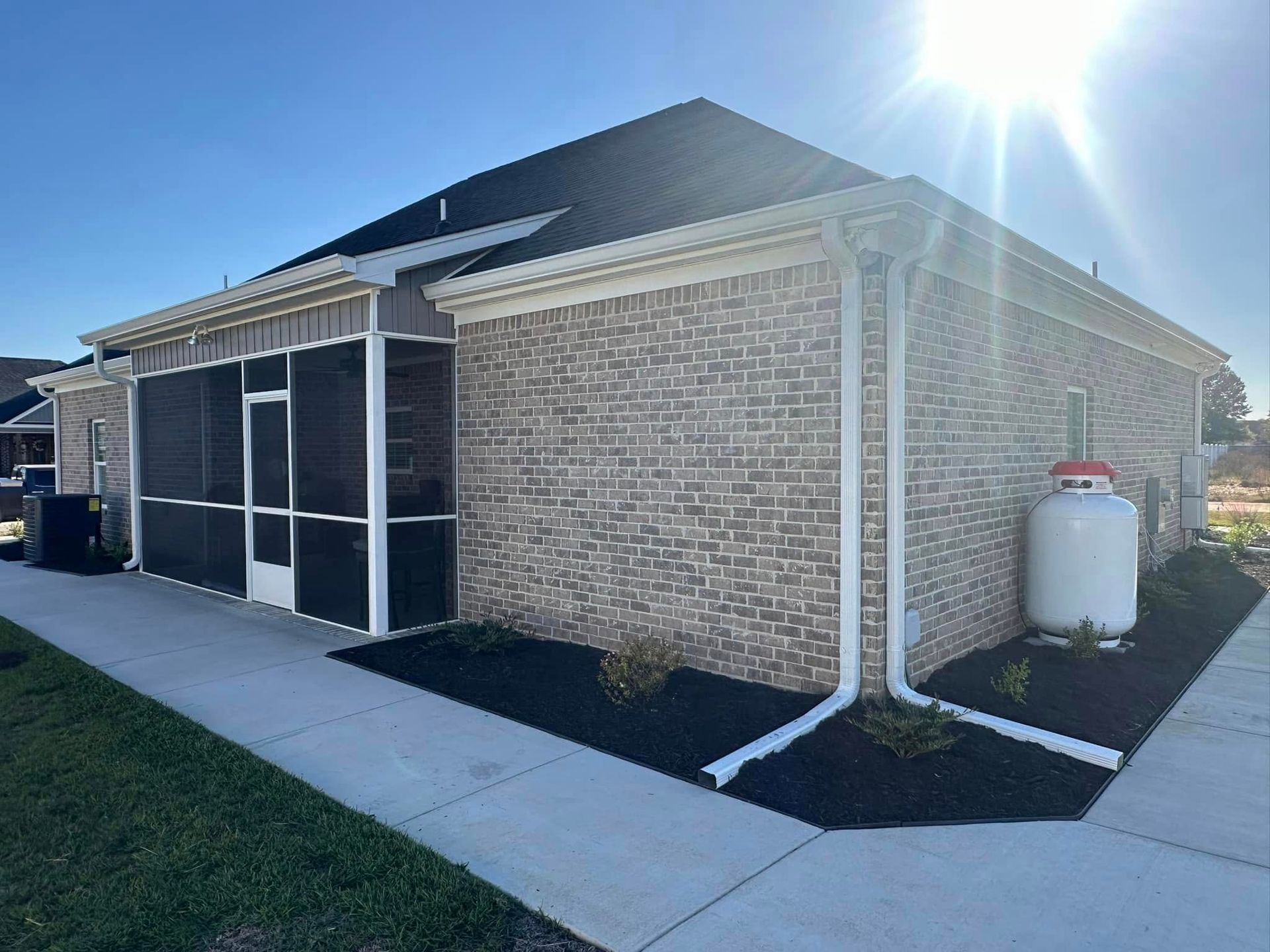 Side view of a brick house with screened porch, concrete walkway, and a white propane tank by the wall.