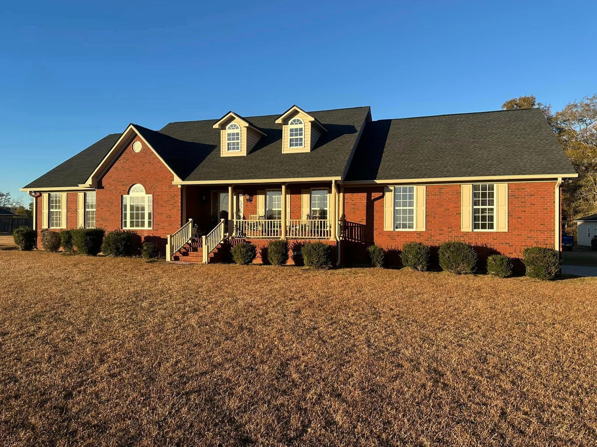 Large red brick suburban house with black roof and front porch, surrounded by a leaf-covered yard under clear sky