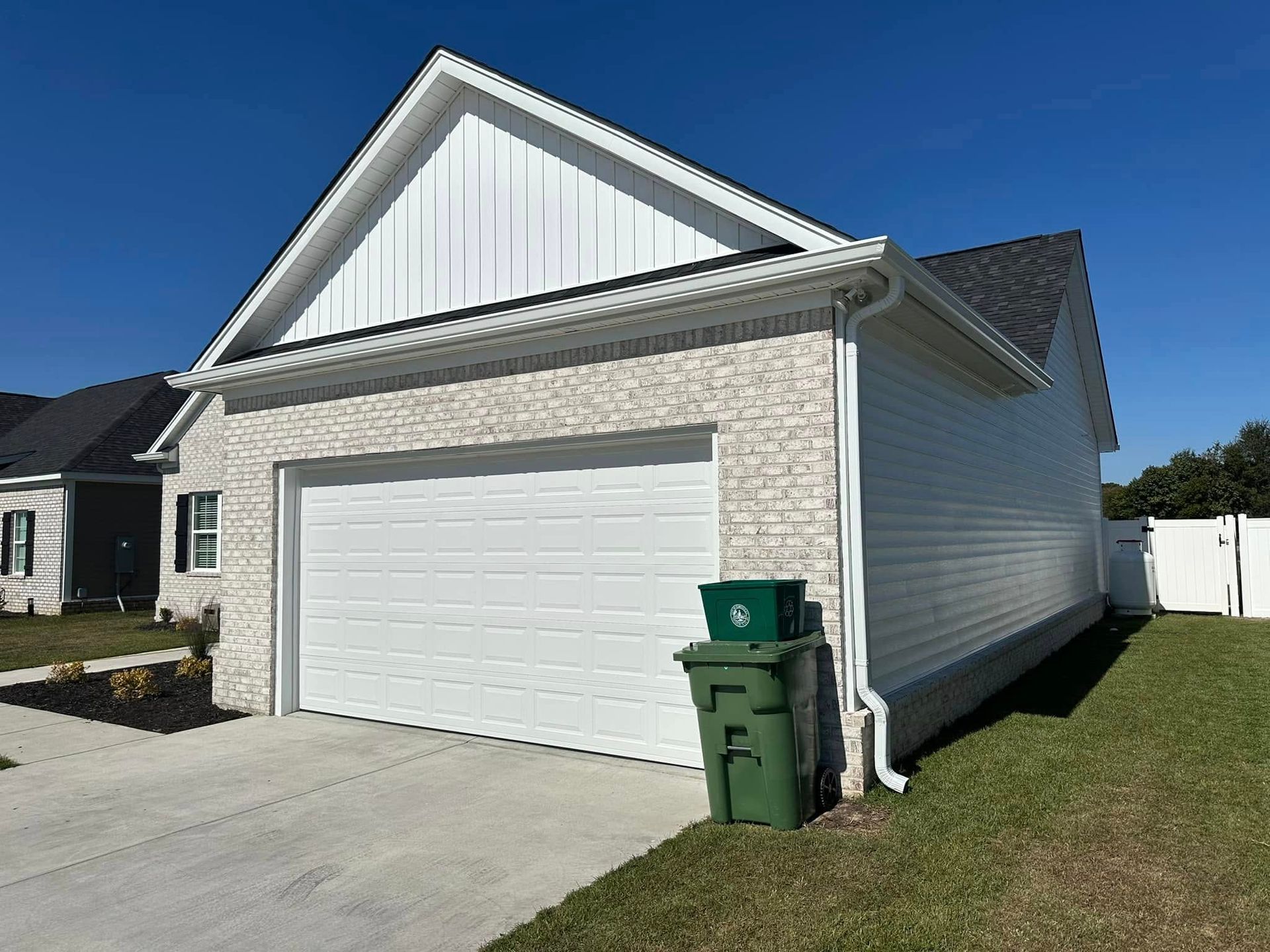 White garage with stone siding and two green trash bins on a driveway beside a grassy lawn