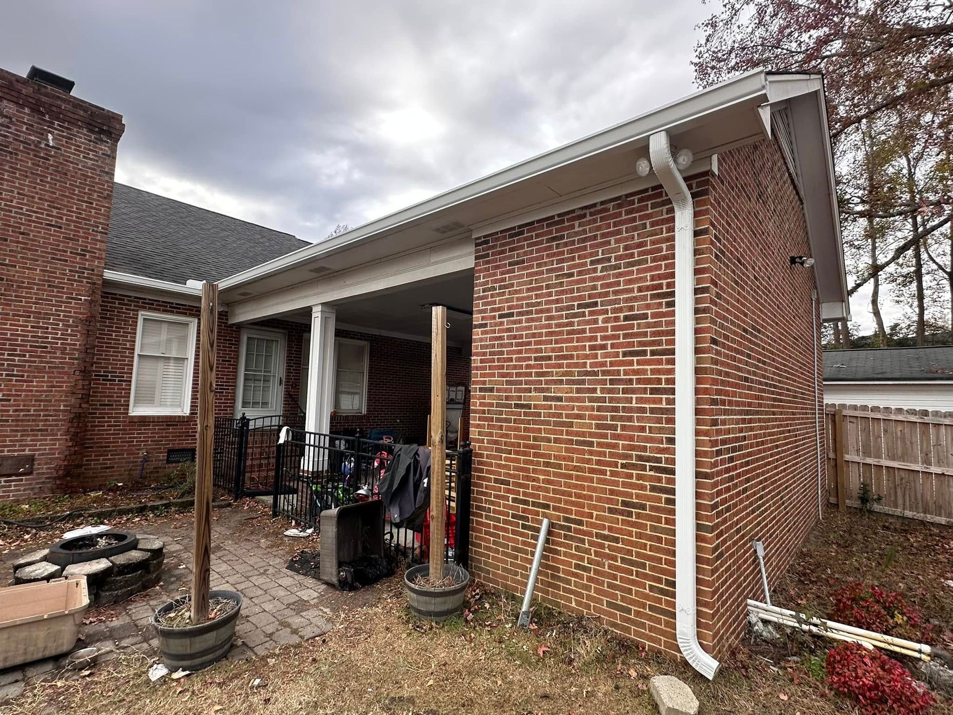 Brick house patio with white gutters, covered porch, and yard tools beside a fenced backyard.