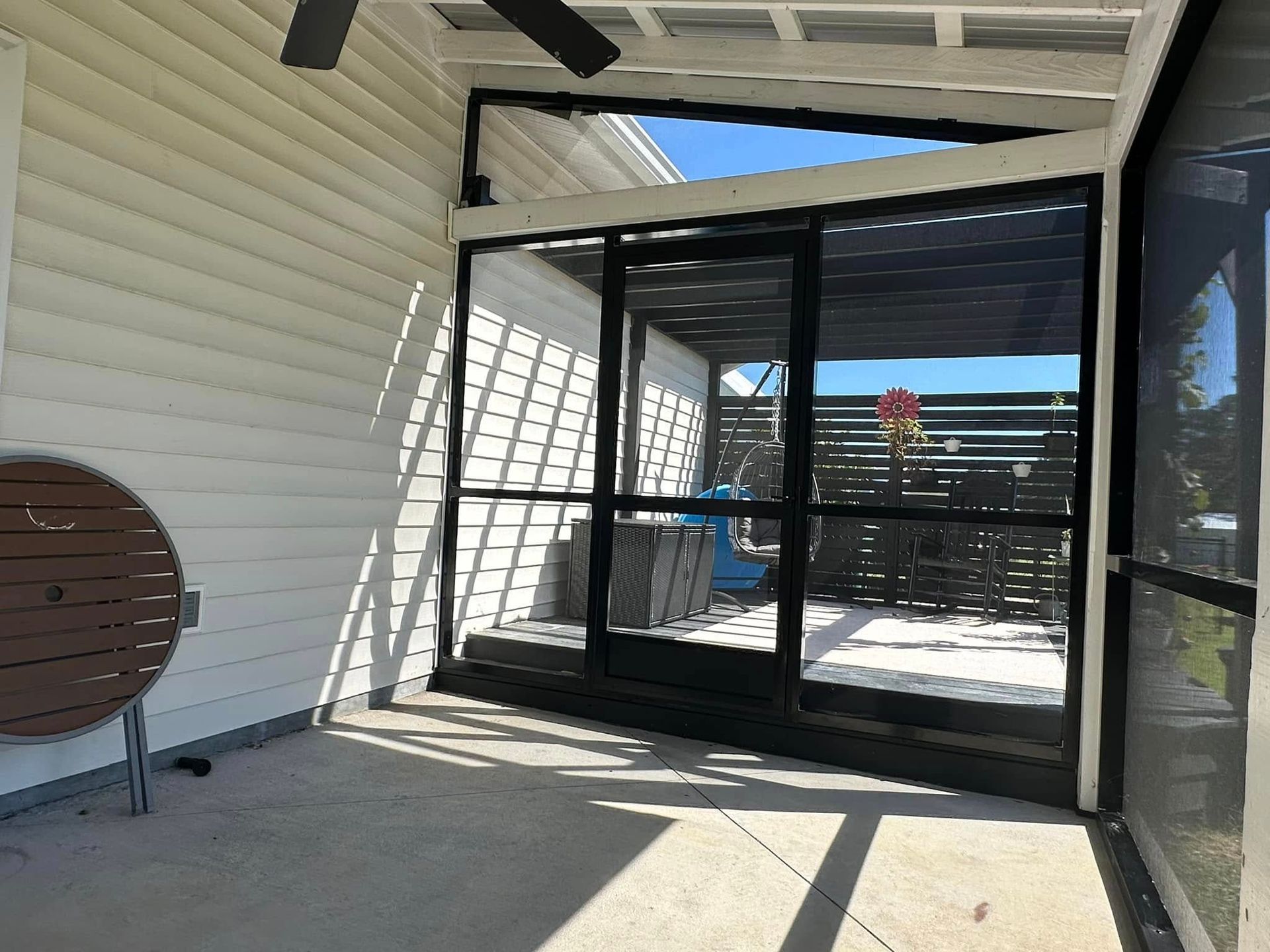 Covered patio with white siding, black-framed glass doors, and outdoor furniture overlooking a sunny deck.
