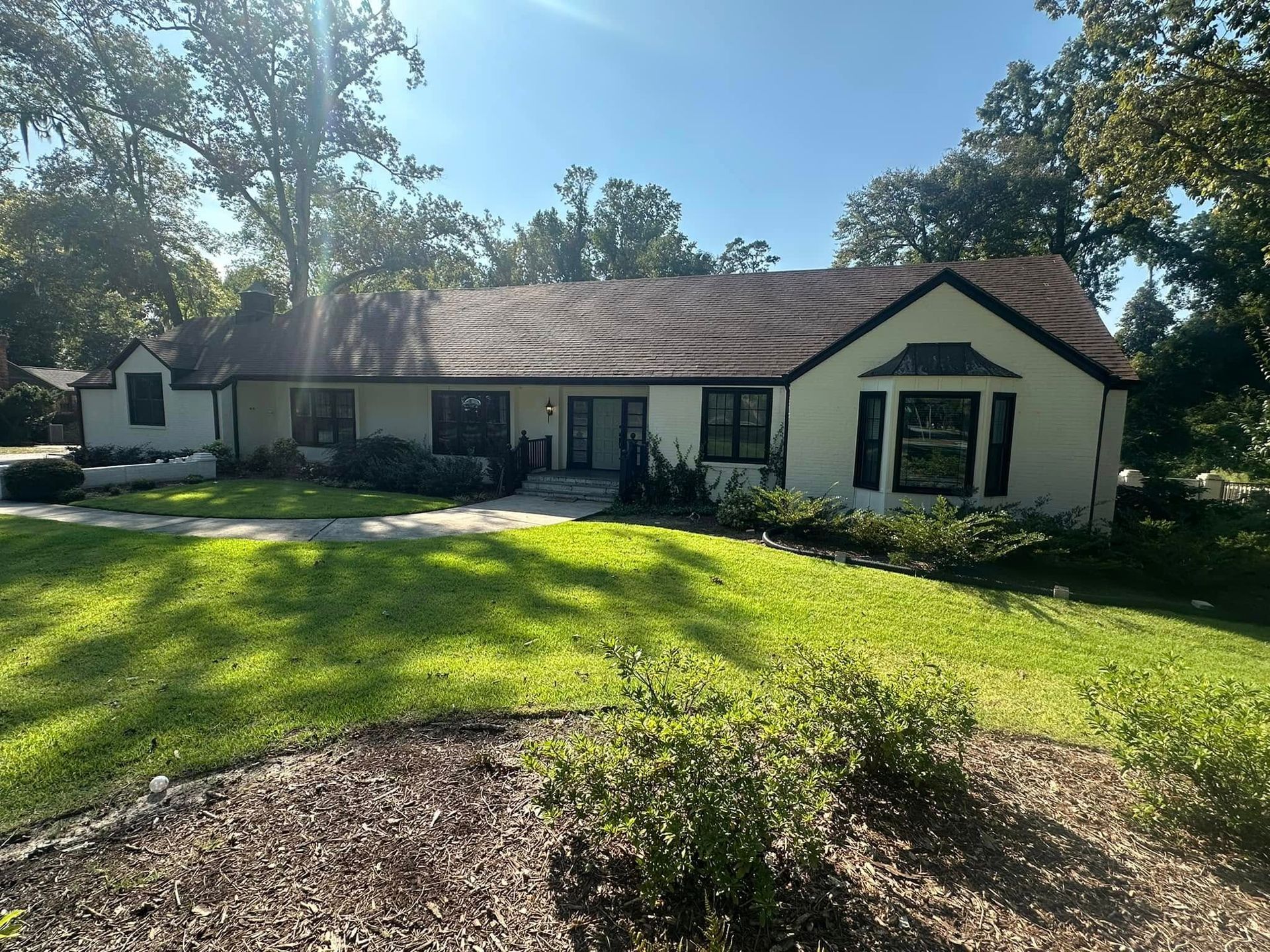 Single-story cream house with a dark roof, front lawn, and trees in bright sunlight