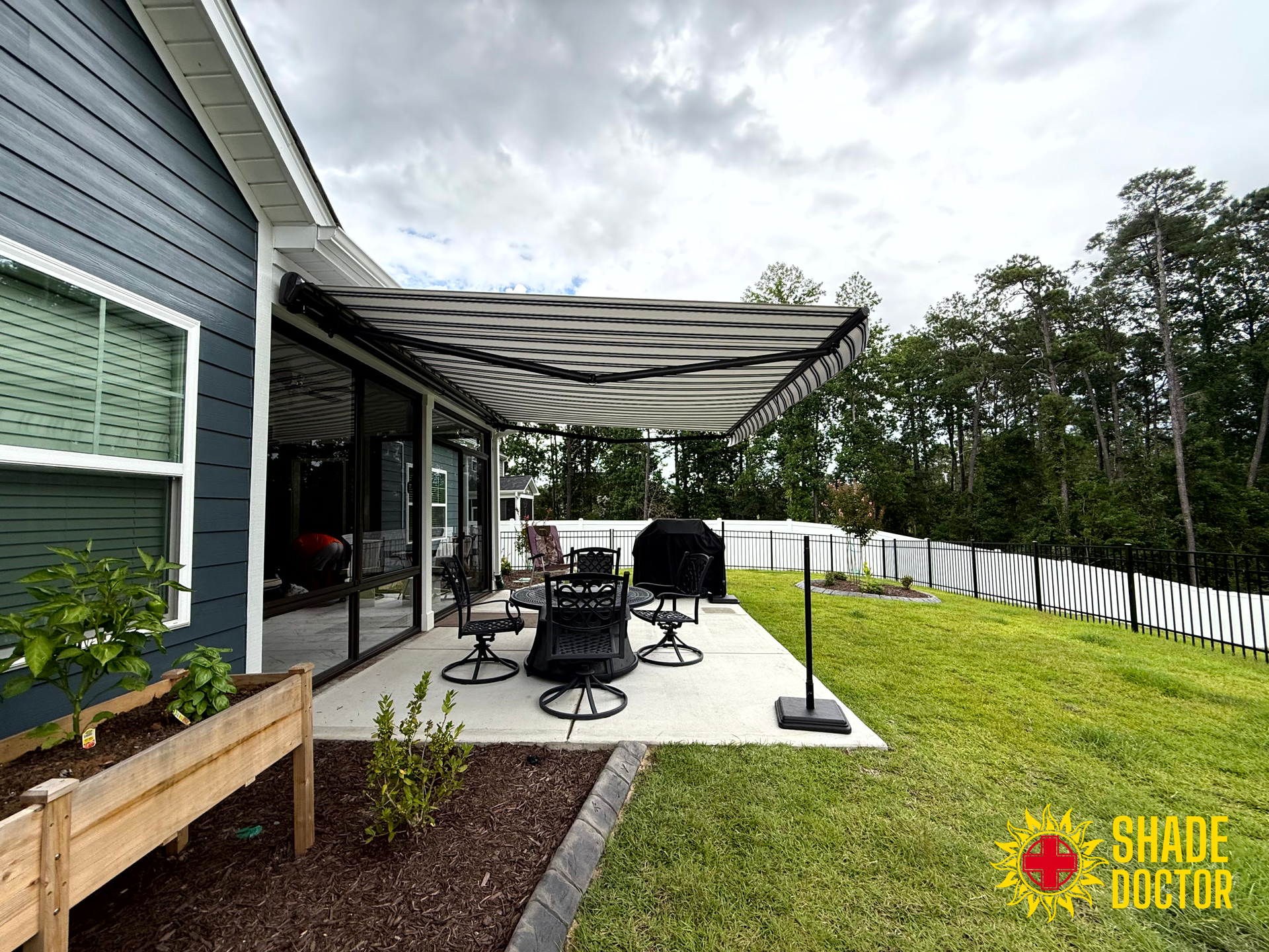 Covered patio with black furniture, grill, and awning beside a house and fenced yard.