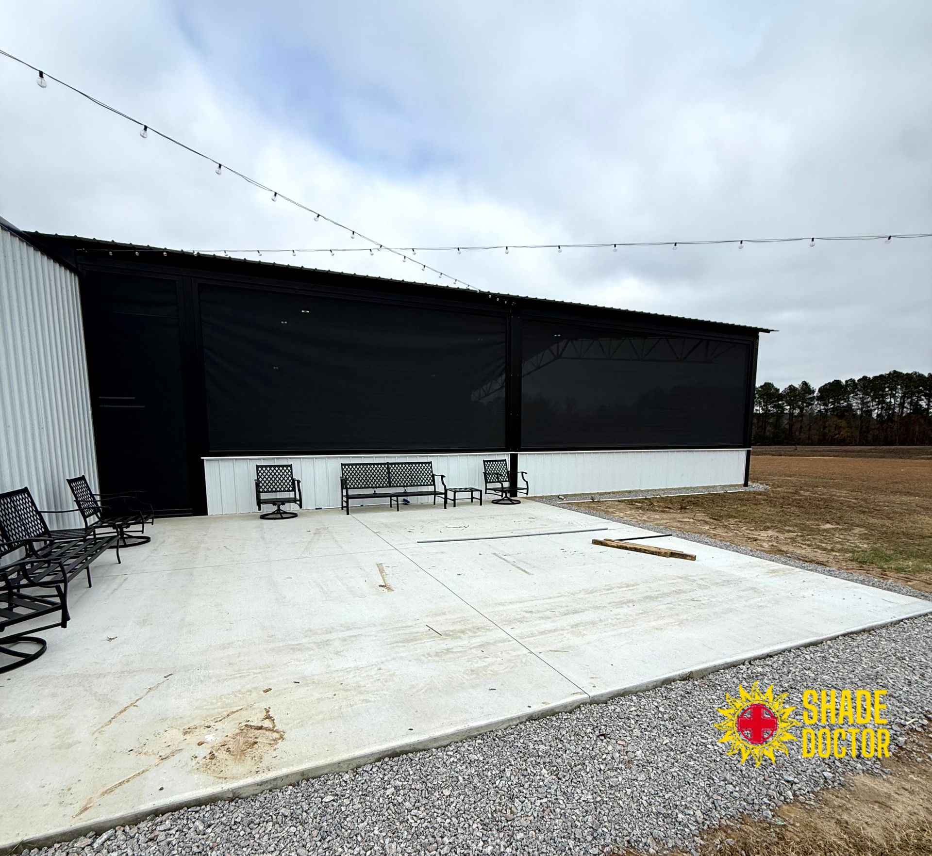 Black pavilion with white concrete patio, string lights, and benches beside an open field.