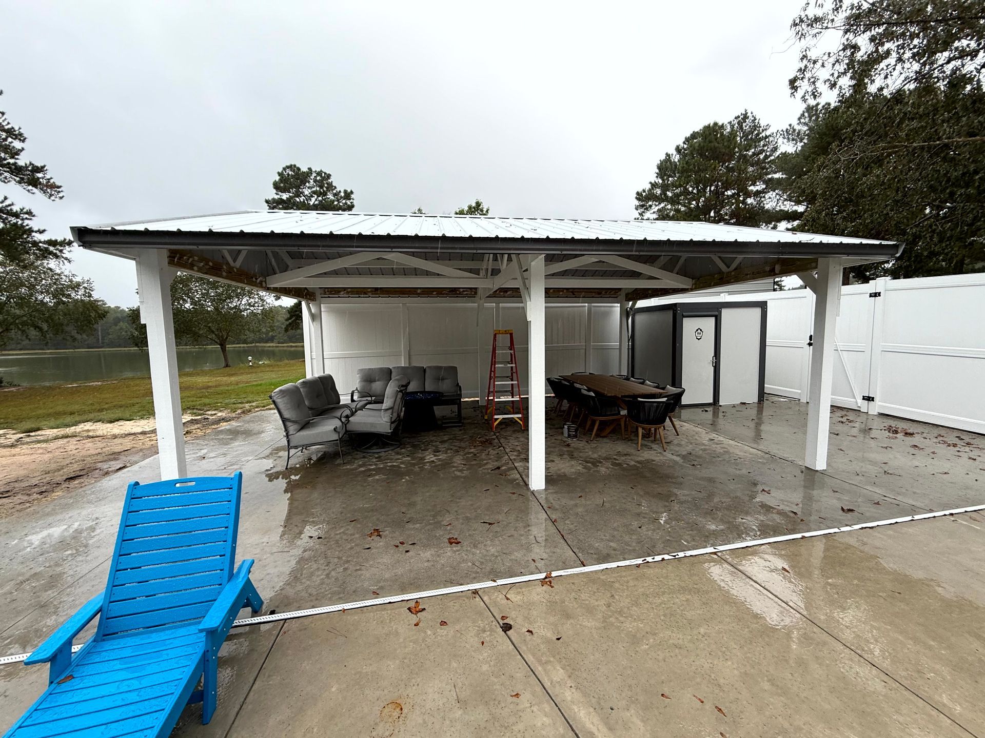 Covered patio with white furniture, ladder, and grill on a wet concrete slab beside a driveway