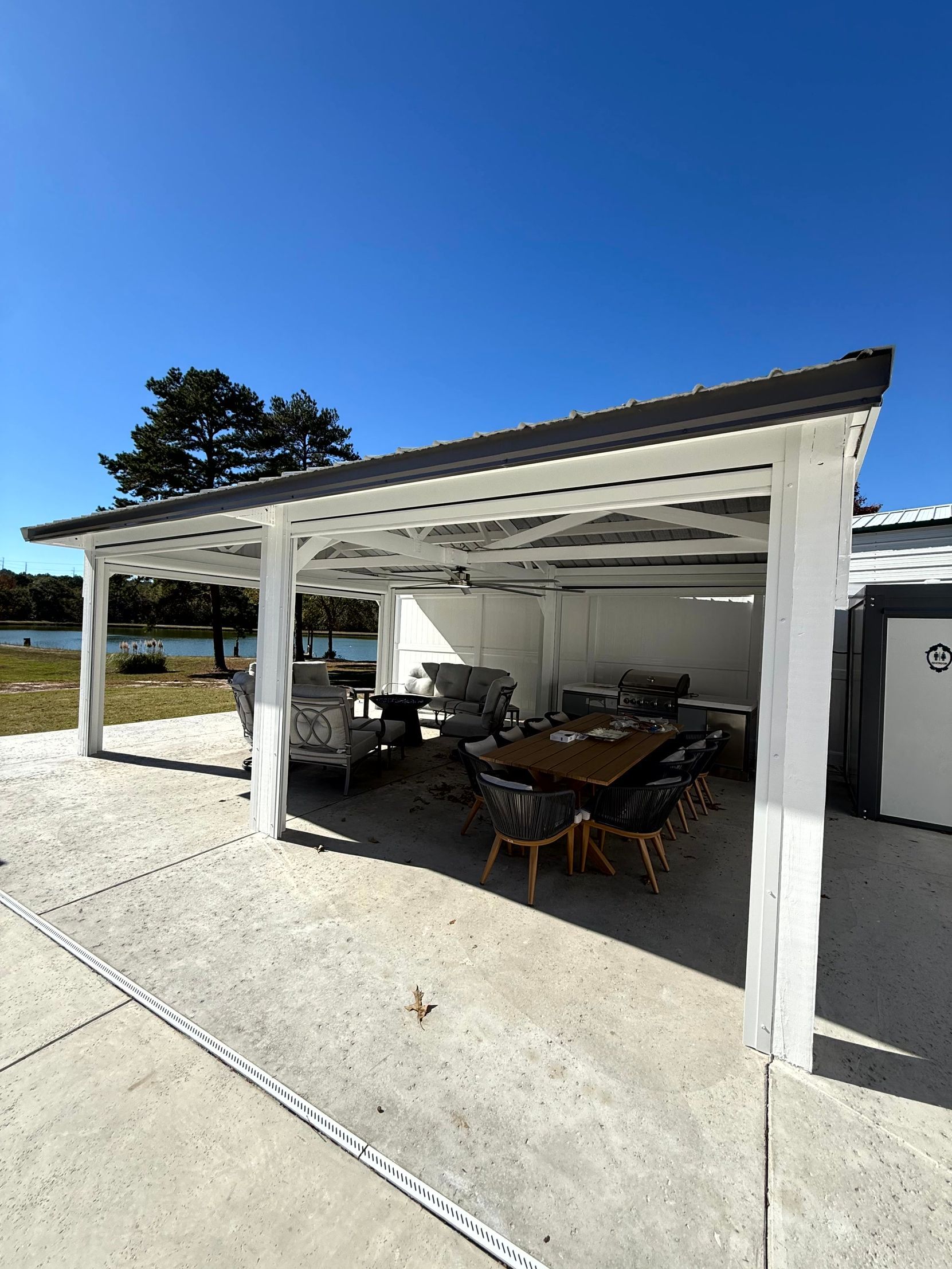 Outdoor patio under a white pergola with table, chairs, and kitchen area on a concrete deck