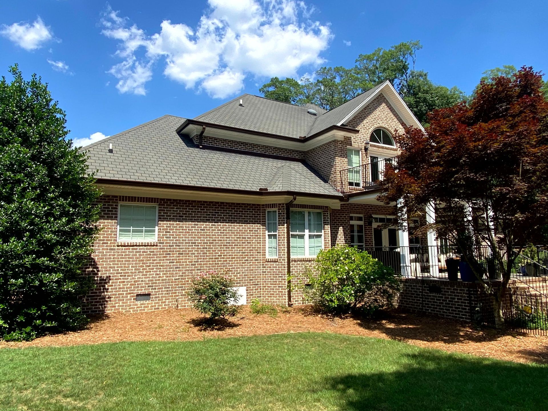 Brick house with gray shingle roof, front porch, and landscaped yard under a clear blue sky