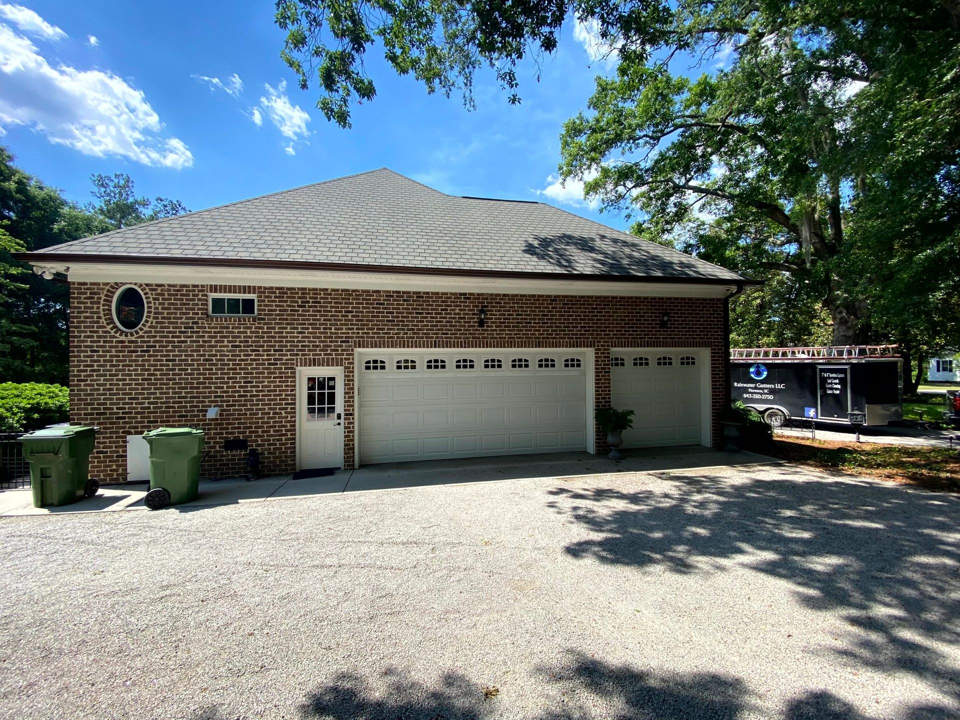 Brick house with a two-car garage, gravel driveway, and green trash bins under a blue sky