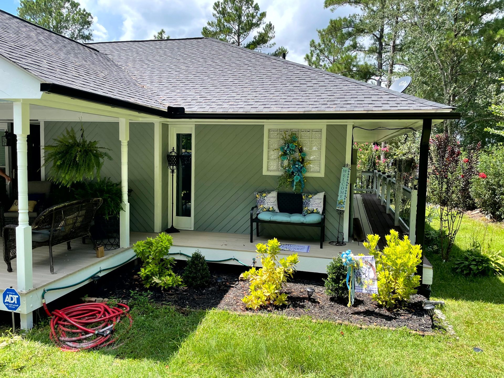 Front porch of a light green house with potted plants, flowers, and a red hose on the lawn