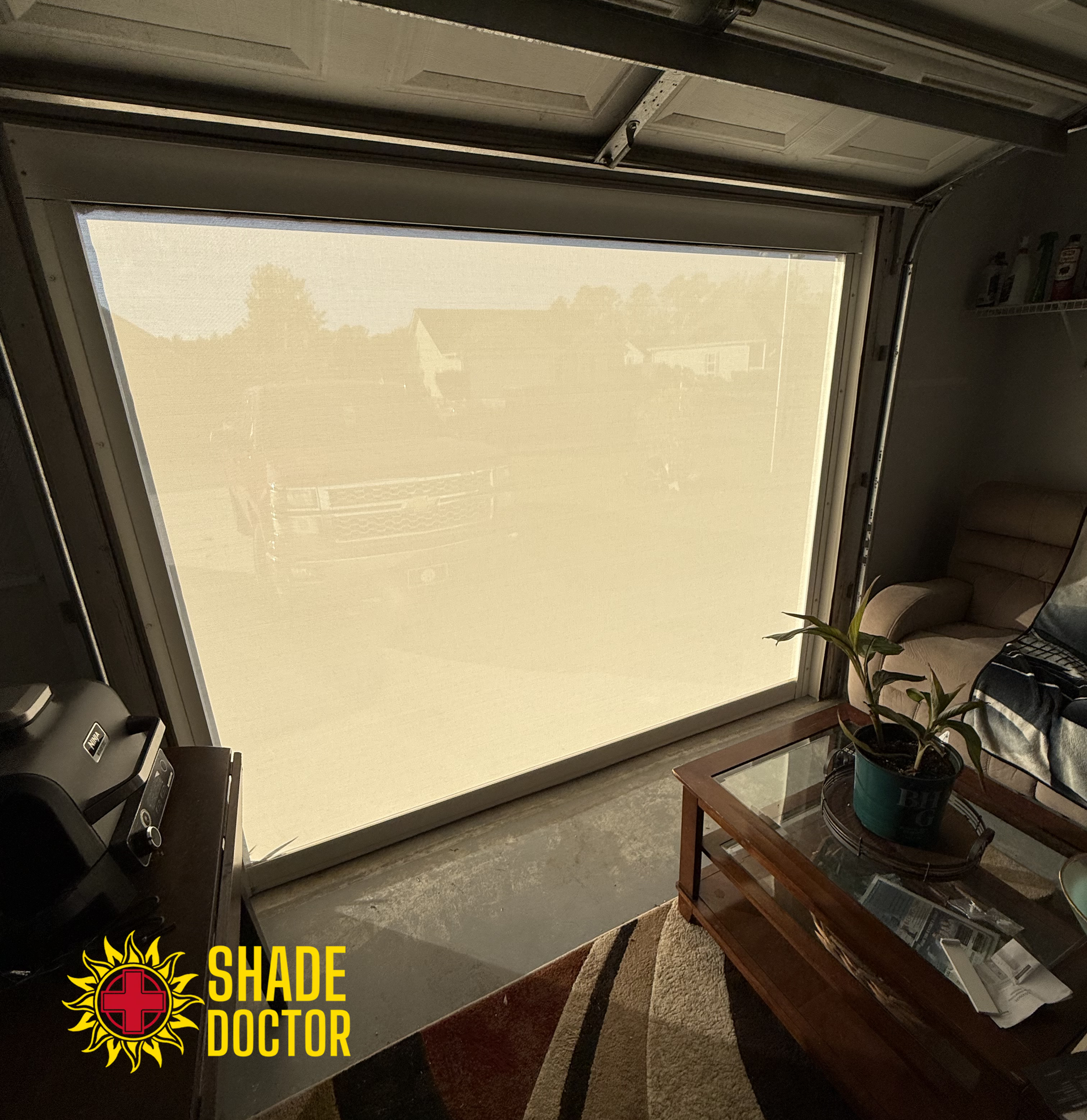 Closed beige roller shade covering a large window in a room with a coffee table and potted plant