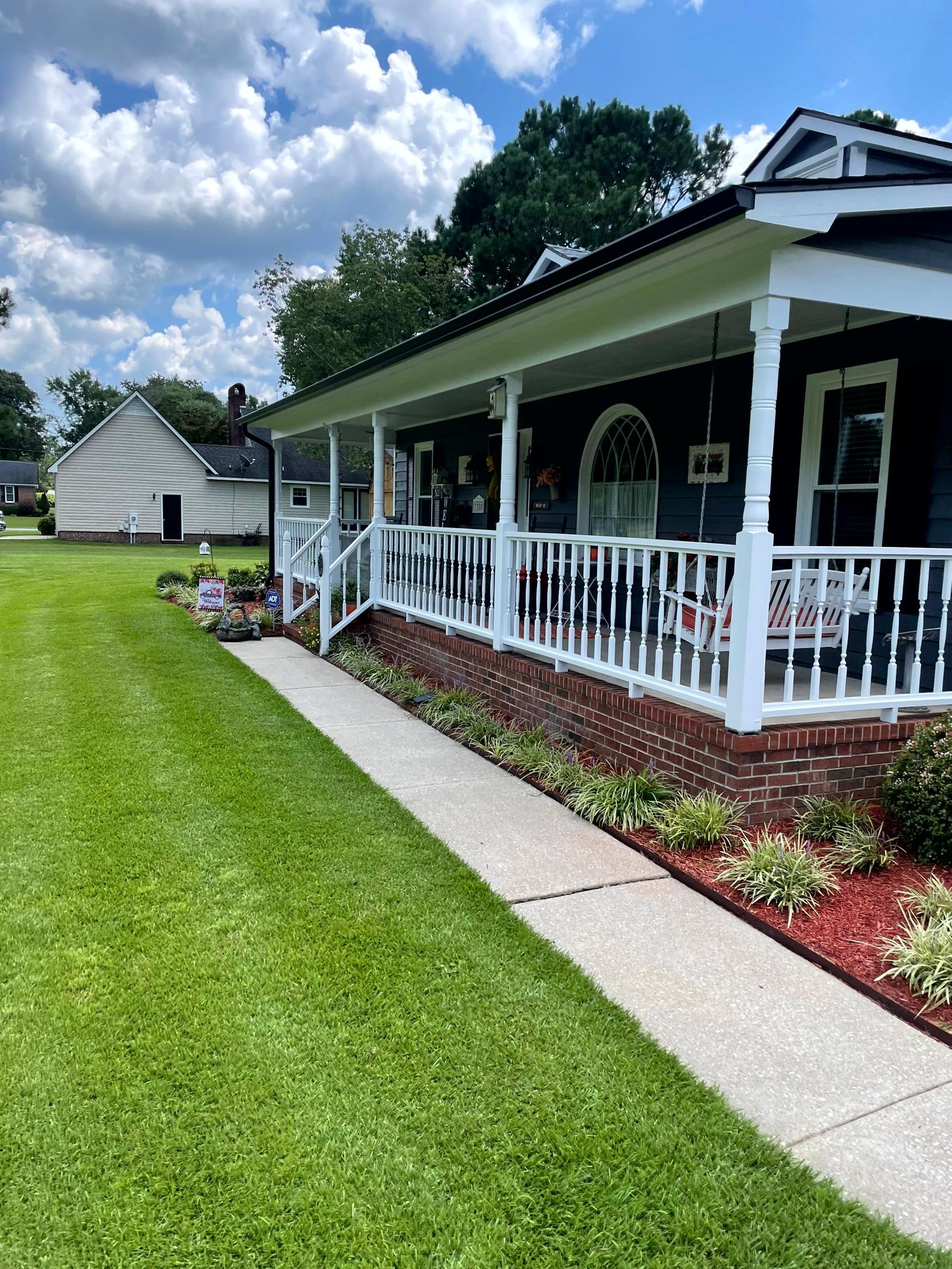 Single-story house with white porch railing, concrete walkway, and green lawn under a cloudy sky