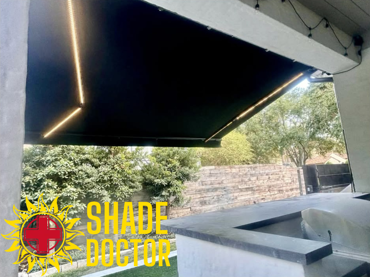 Black motorized patio shade extended over a poolside patio, with trees and stone wall beyond