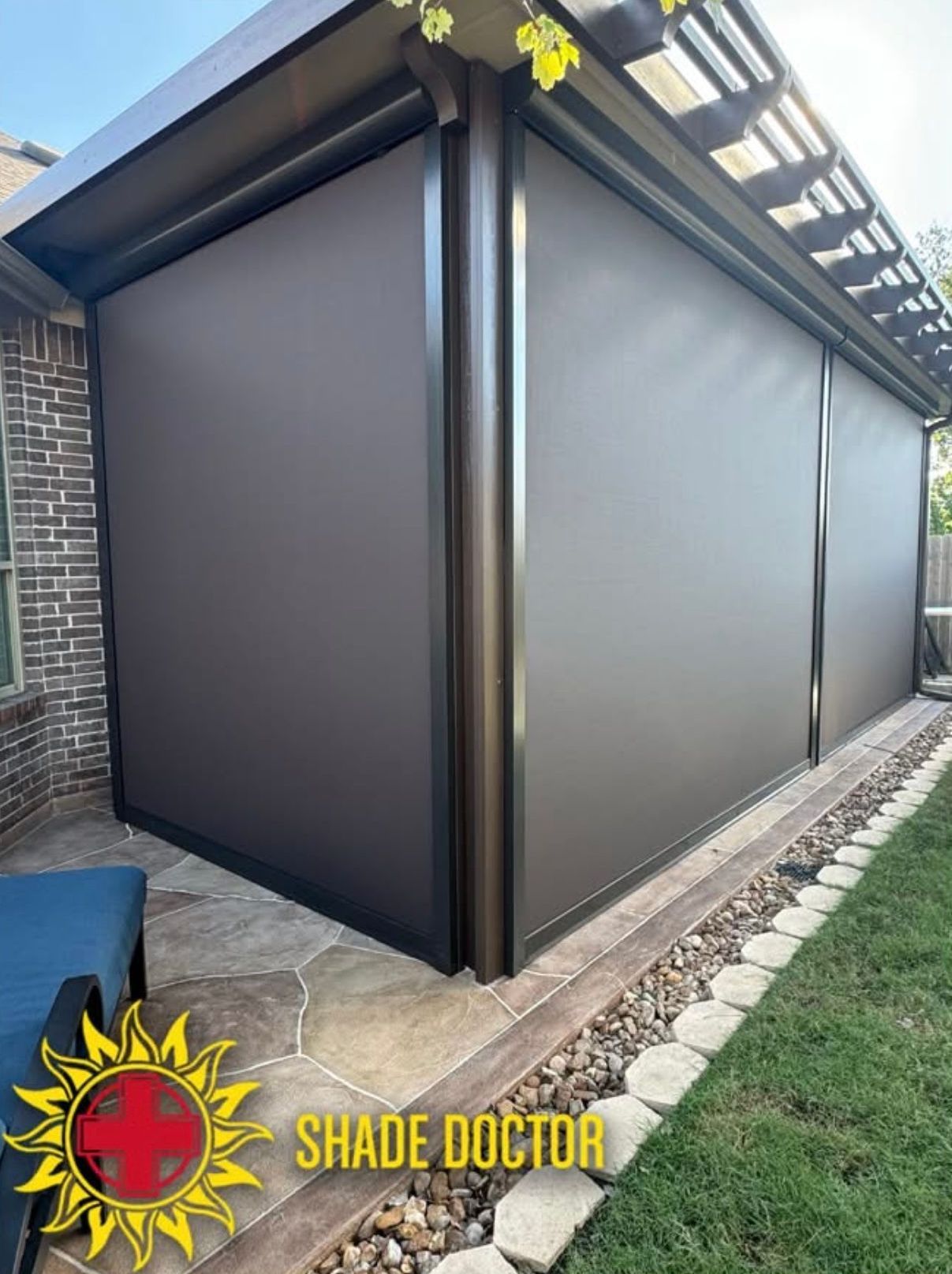 Outdoor patio with dark brown retractable shade screens under a pergola beside a lawn and brick wall