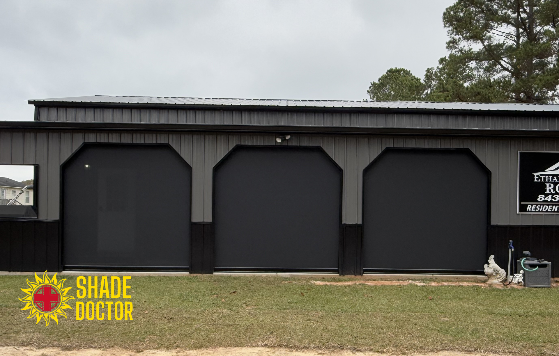 Black building with three dark garage doors and Shade Doctor signs on the front lawn.