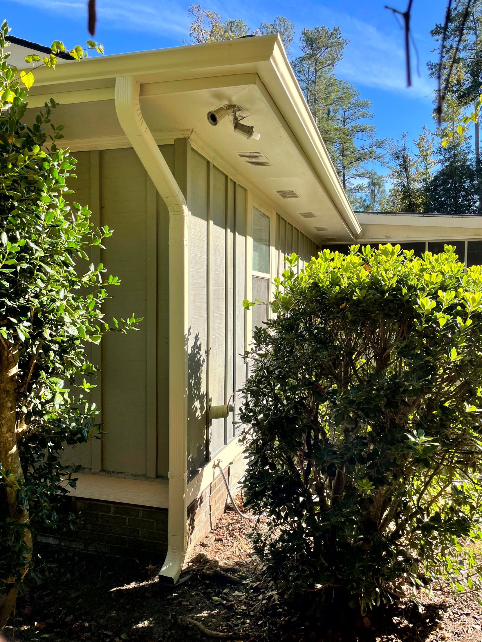 Small beige house exterior with white trim, bushes, and a bright blue sky.