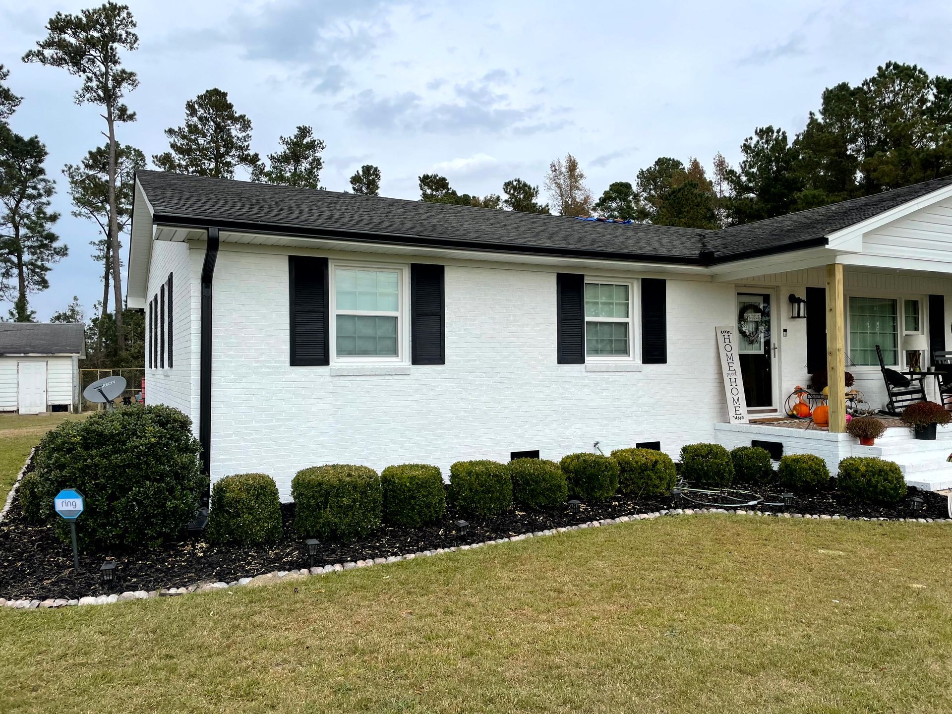 Single-story white house with black shutters, front porch, and neatly trimmed hedges under a cloudy sky