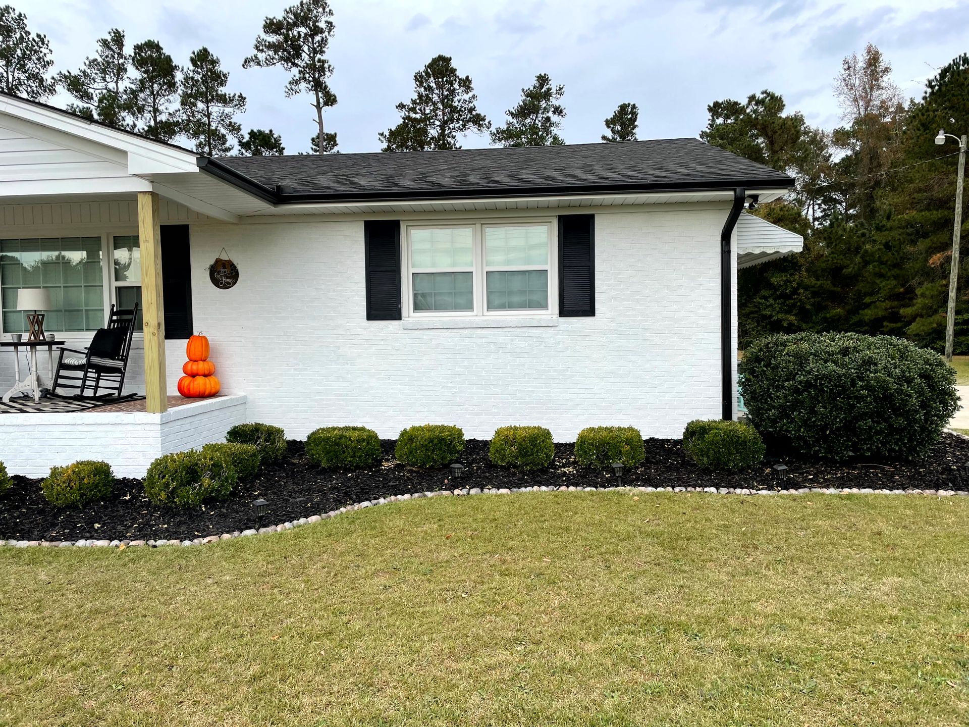 White house exterior with black shutters, trimmed shrubs, and a small porch with an orange pumpkin decoration.