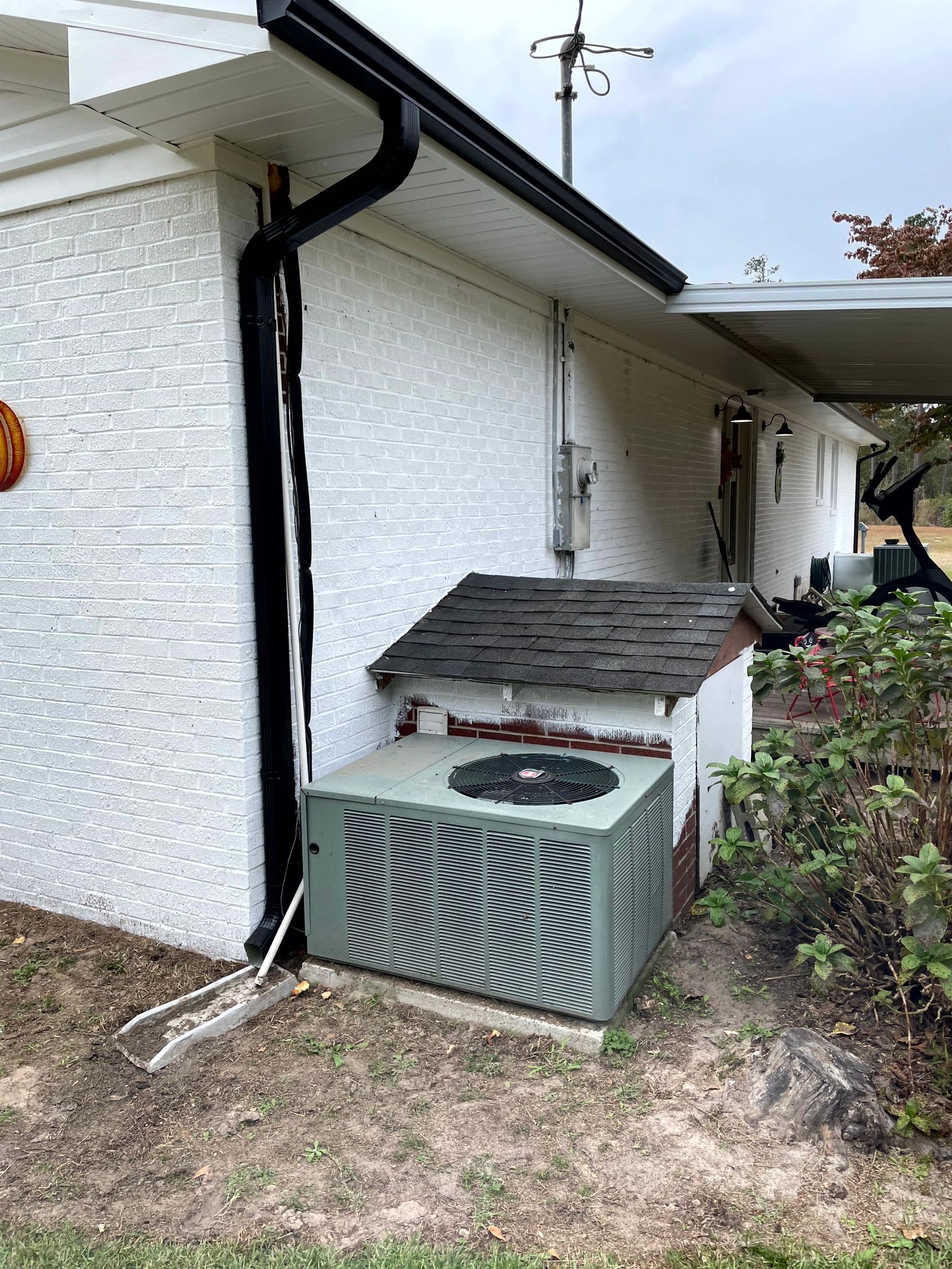 Outdoor HVAC unit beside a white house, partially covered by a small wooden roof.