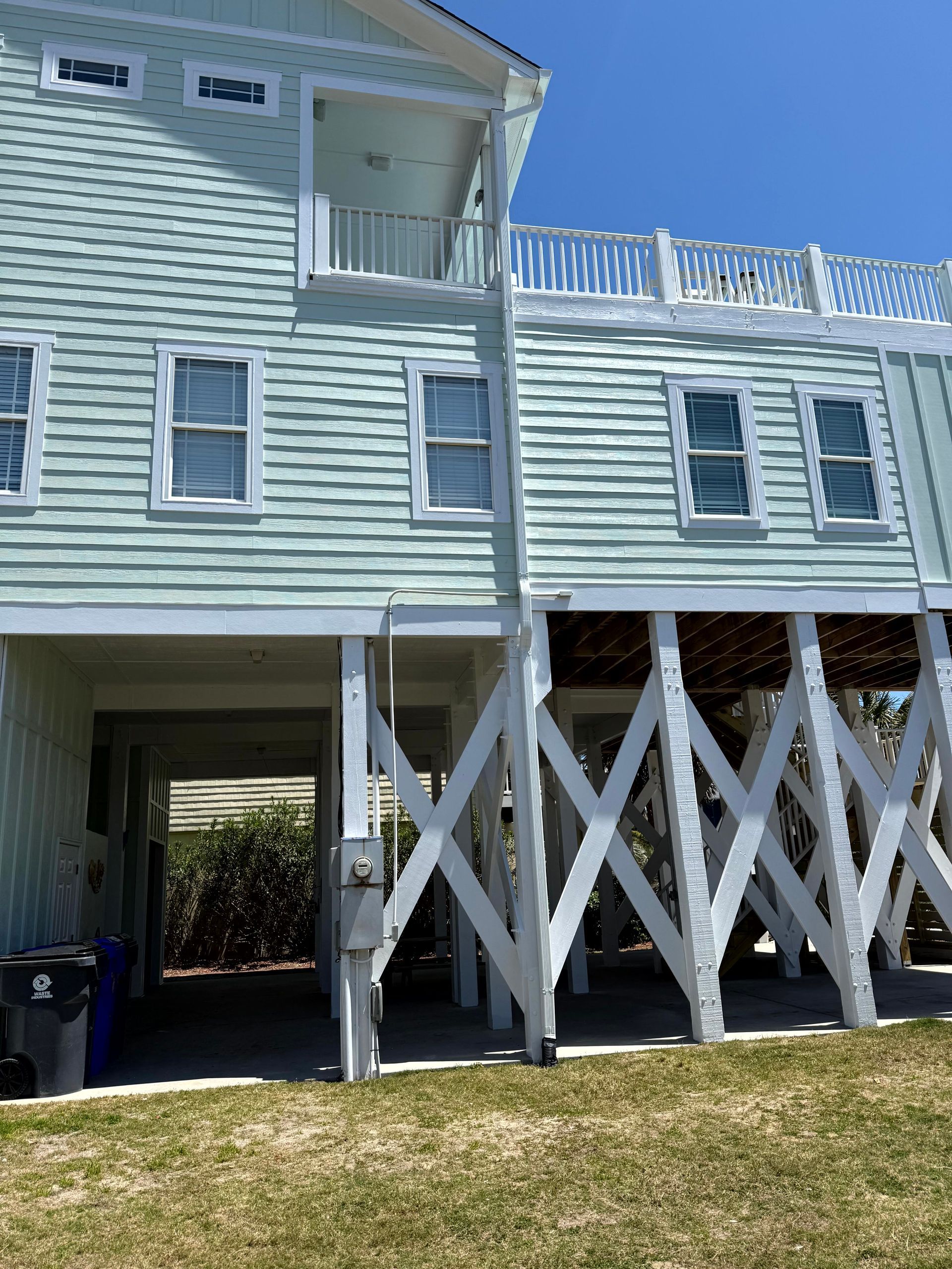 Light blue coastal house on stilts with white crisscross supports and shaded parking area beneath