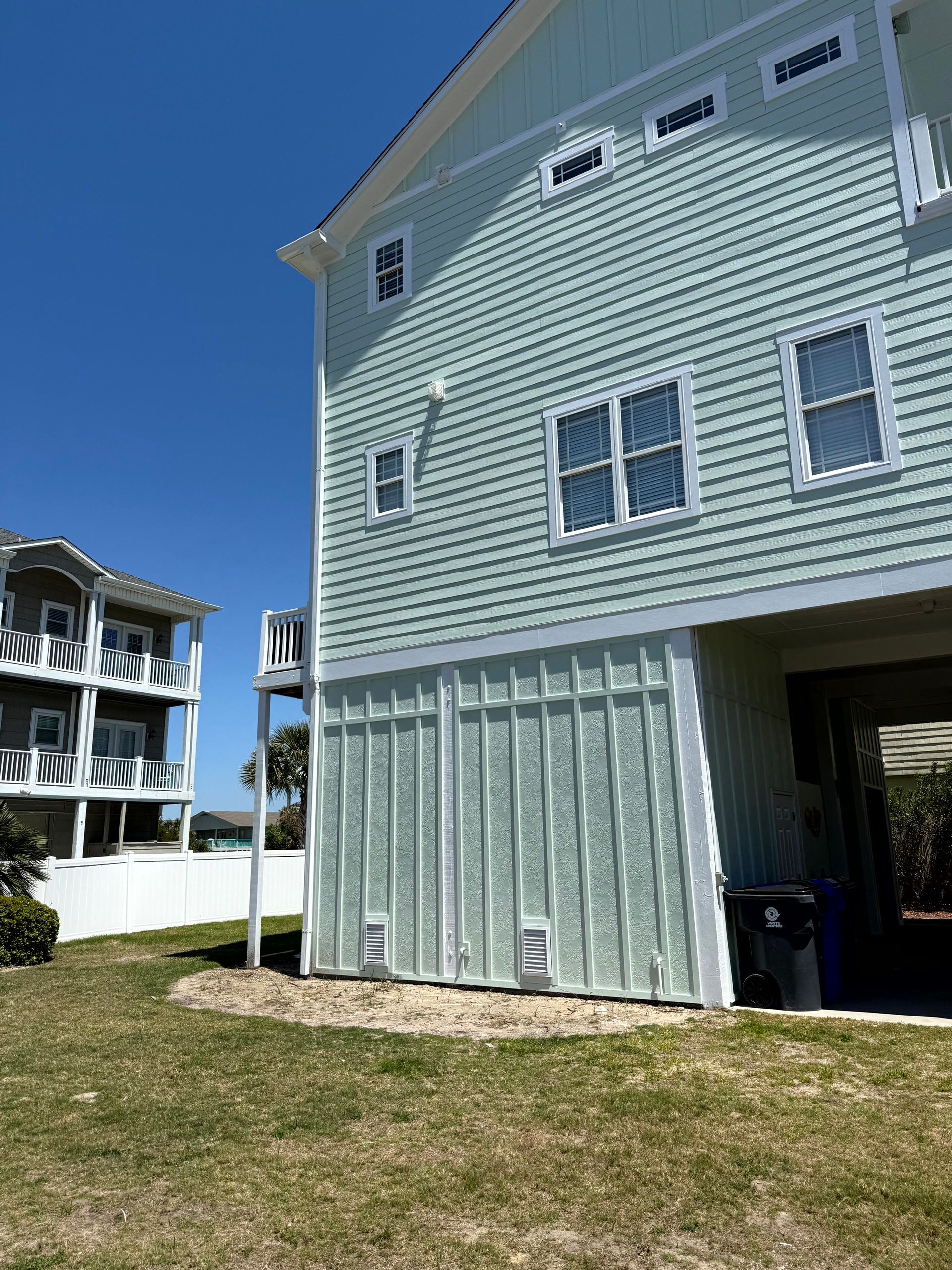 Light blue beach house with white trim and covered ground level beside a grassy yard under a clear sky