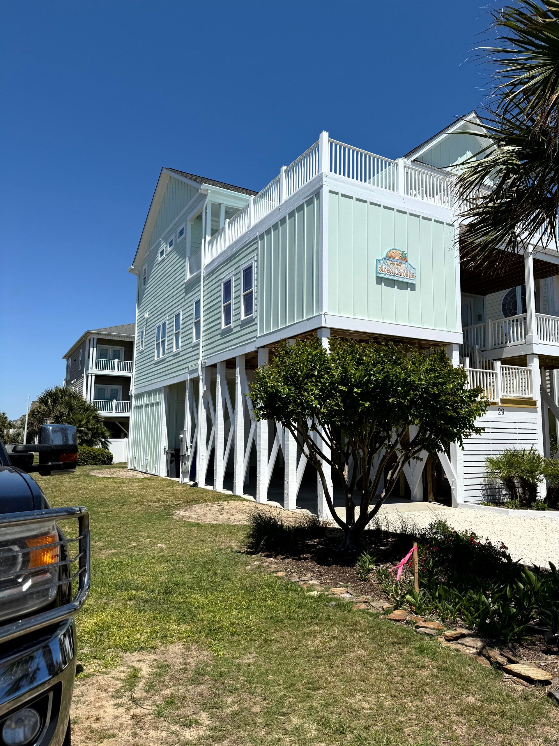 Pastel seaside house on stilts with white trim and a green yard under a clear blue sky