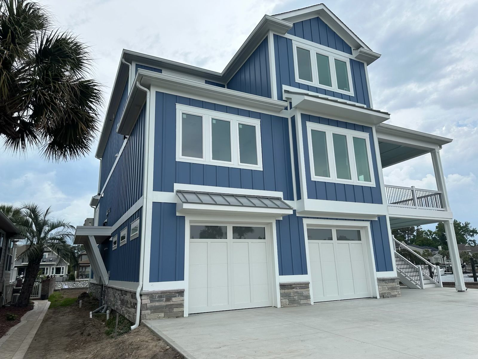 Blue beach house with white trim, balconies, and two garage doors on a palm-lined driveway