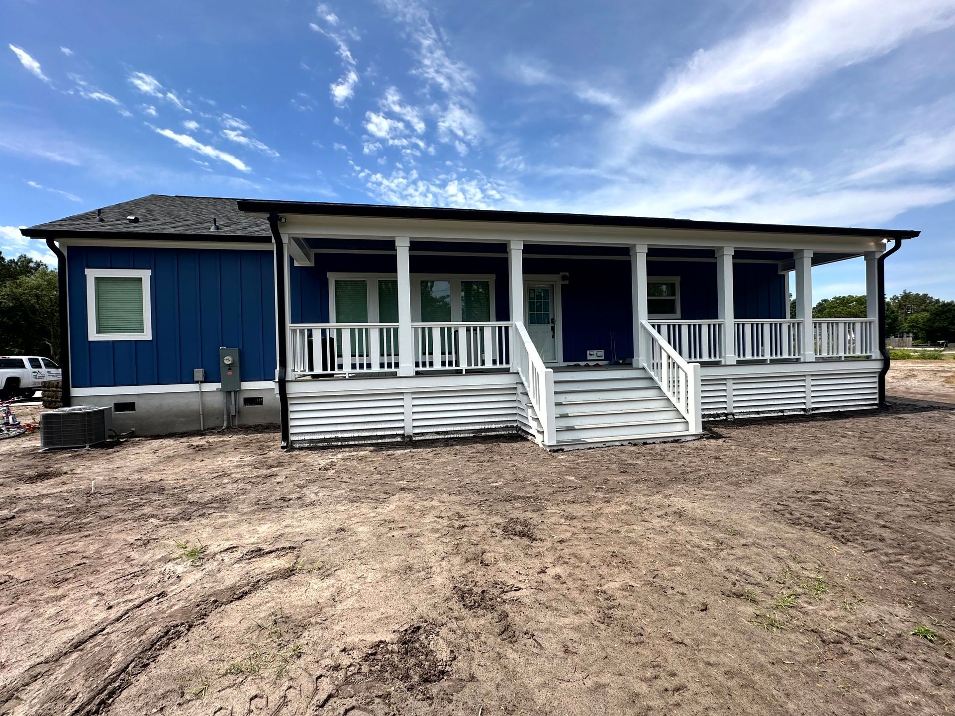 Blue house with white porch railings on a dirt lot under a partly cloudy sky