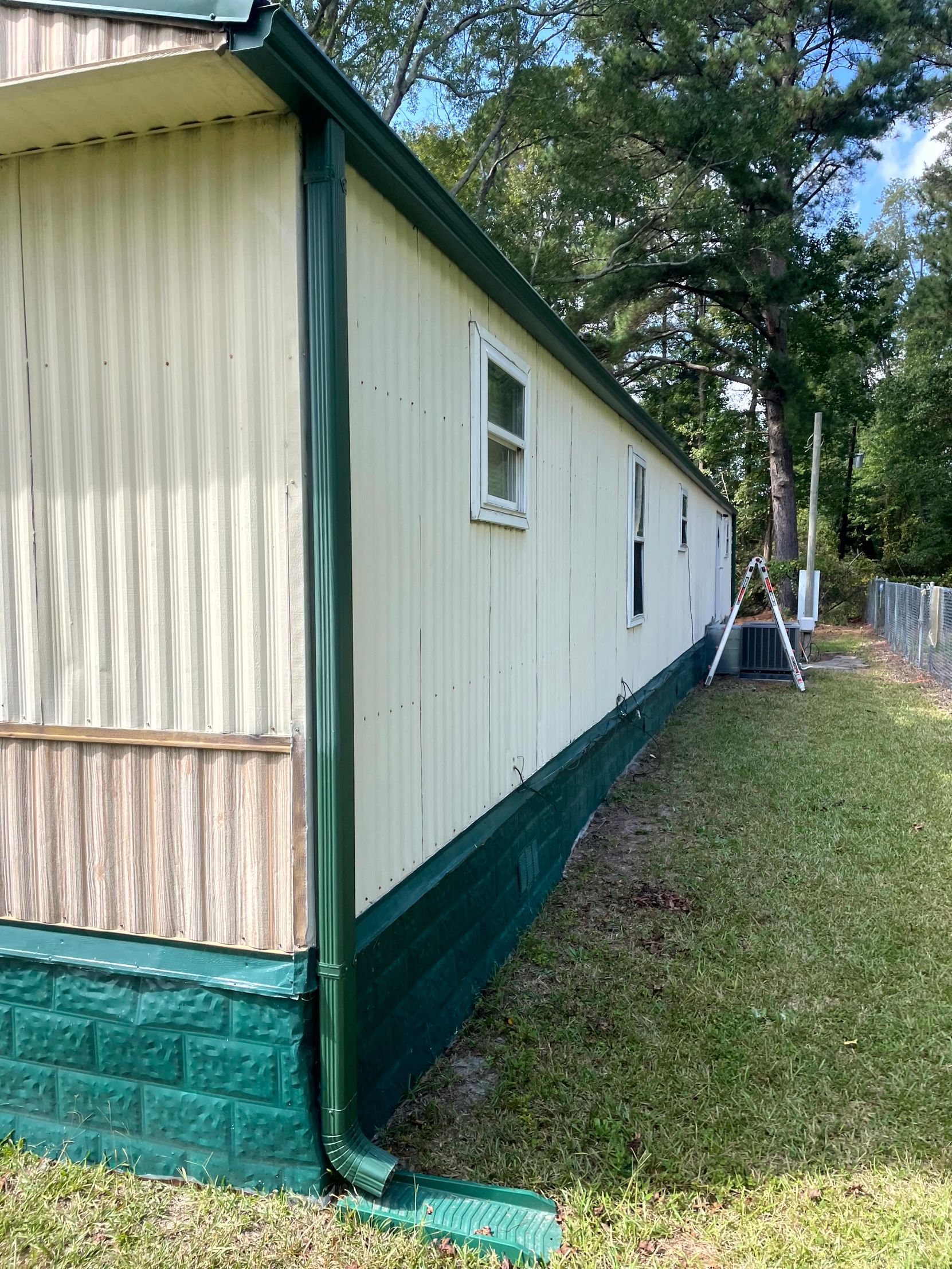 White mobile home with green trim on a grassy lot, viewed from the side under trees