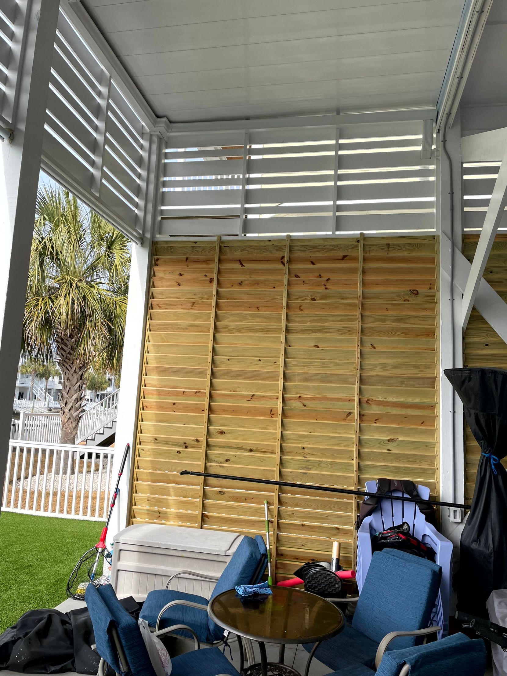 Stacked wooden planks under a metal shelter, with folded chairs and a bag in the foreground.
