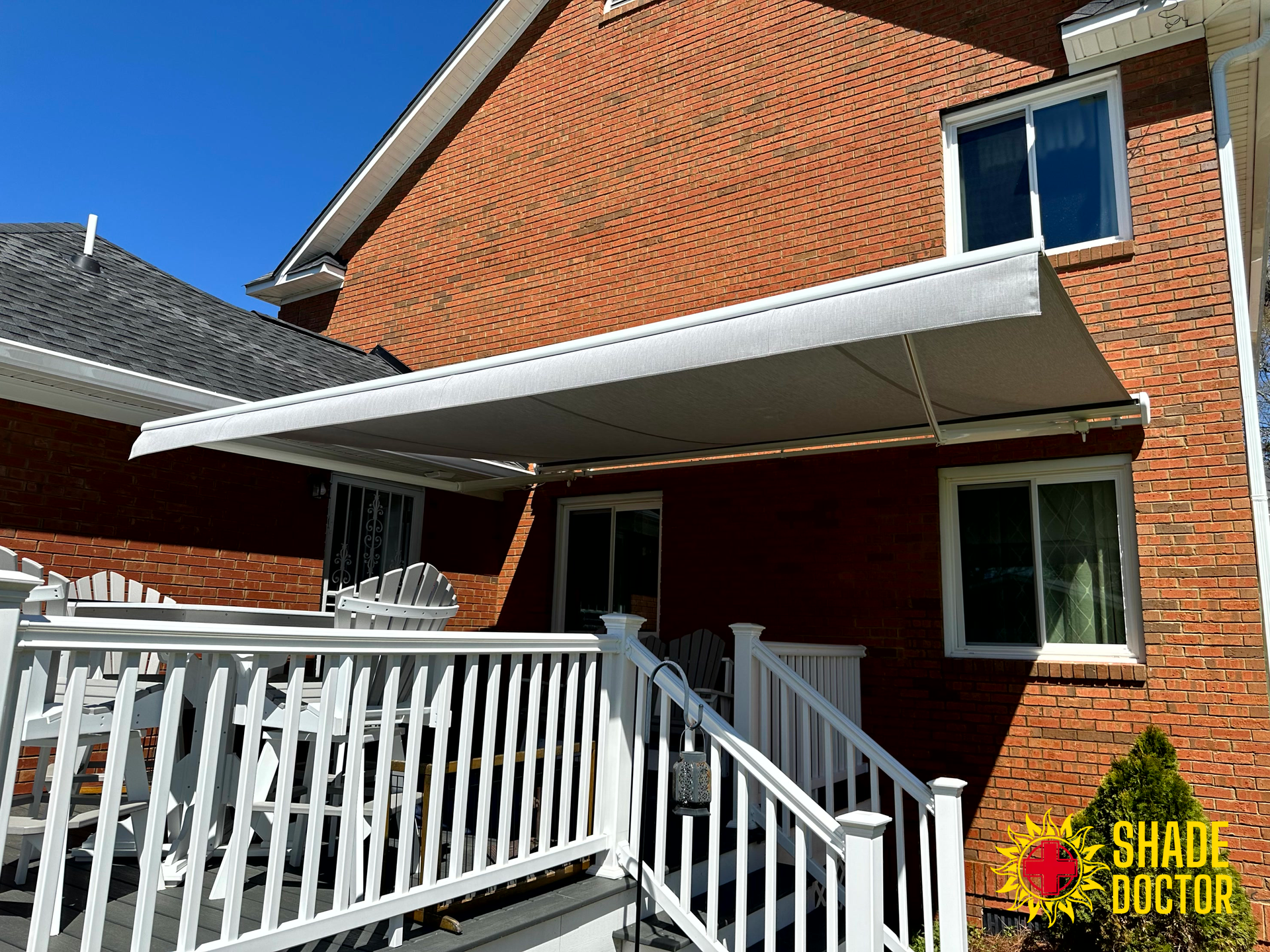Brick house patio with white railing and awning, viewed from a backyard deck.