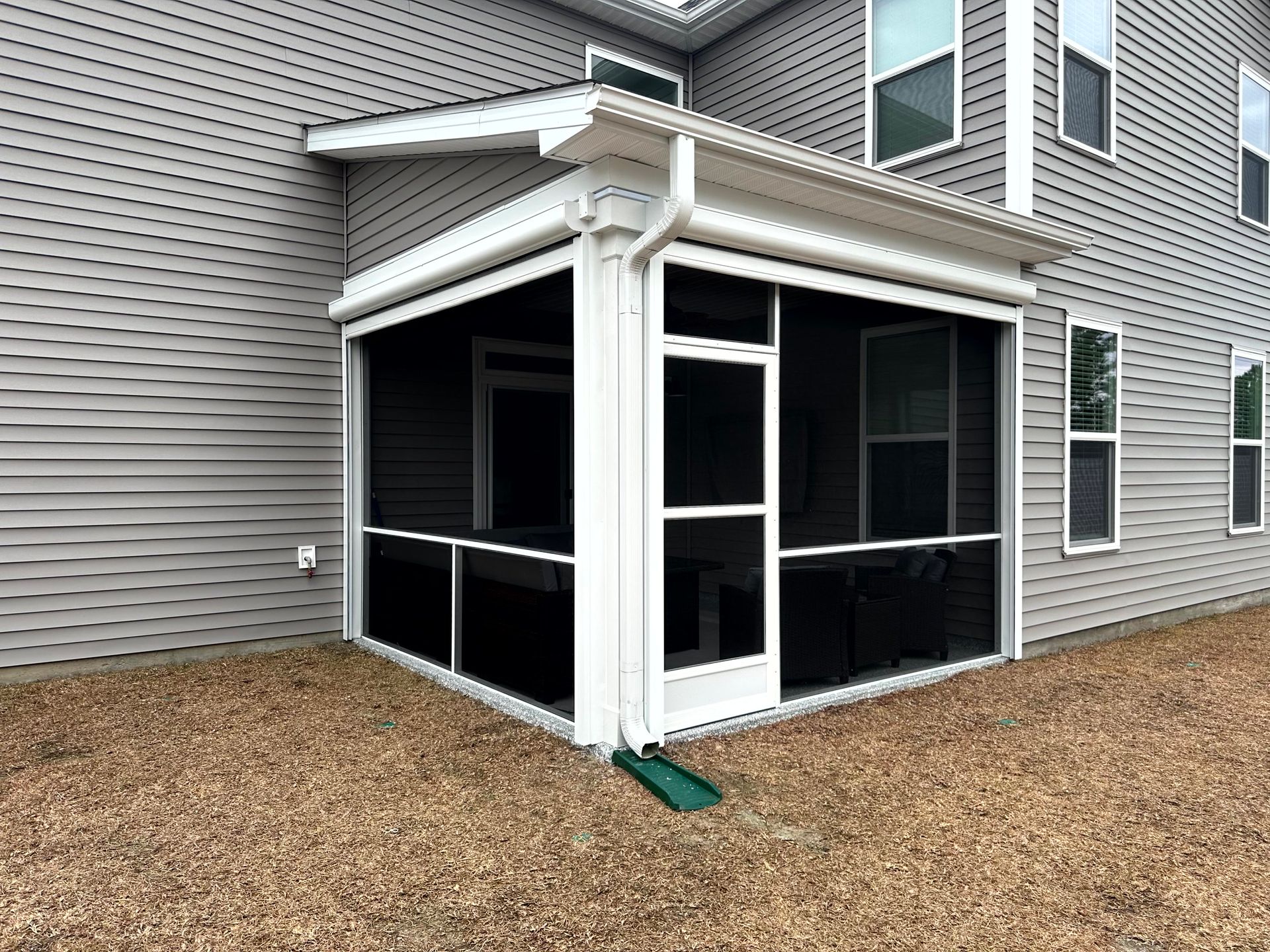Screened porch with white trim attached to a gray-sided house, surrounded by gravel mulch.