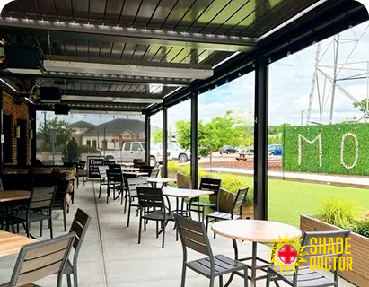 Outdoor café patio with tables and chairs under a roof, overlooking a green lawn