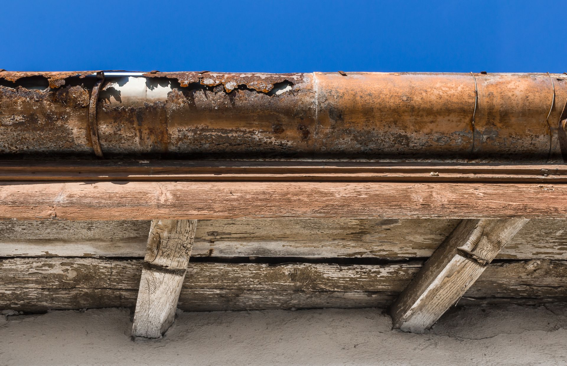 Rusted metal pipes and weathered wooden beams under a roofline against a concrete wall