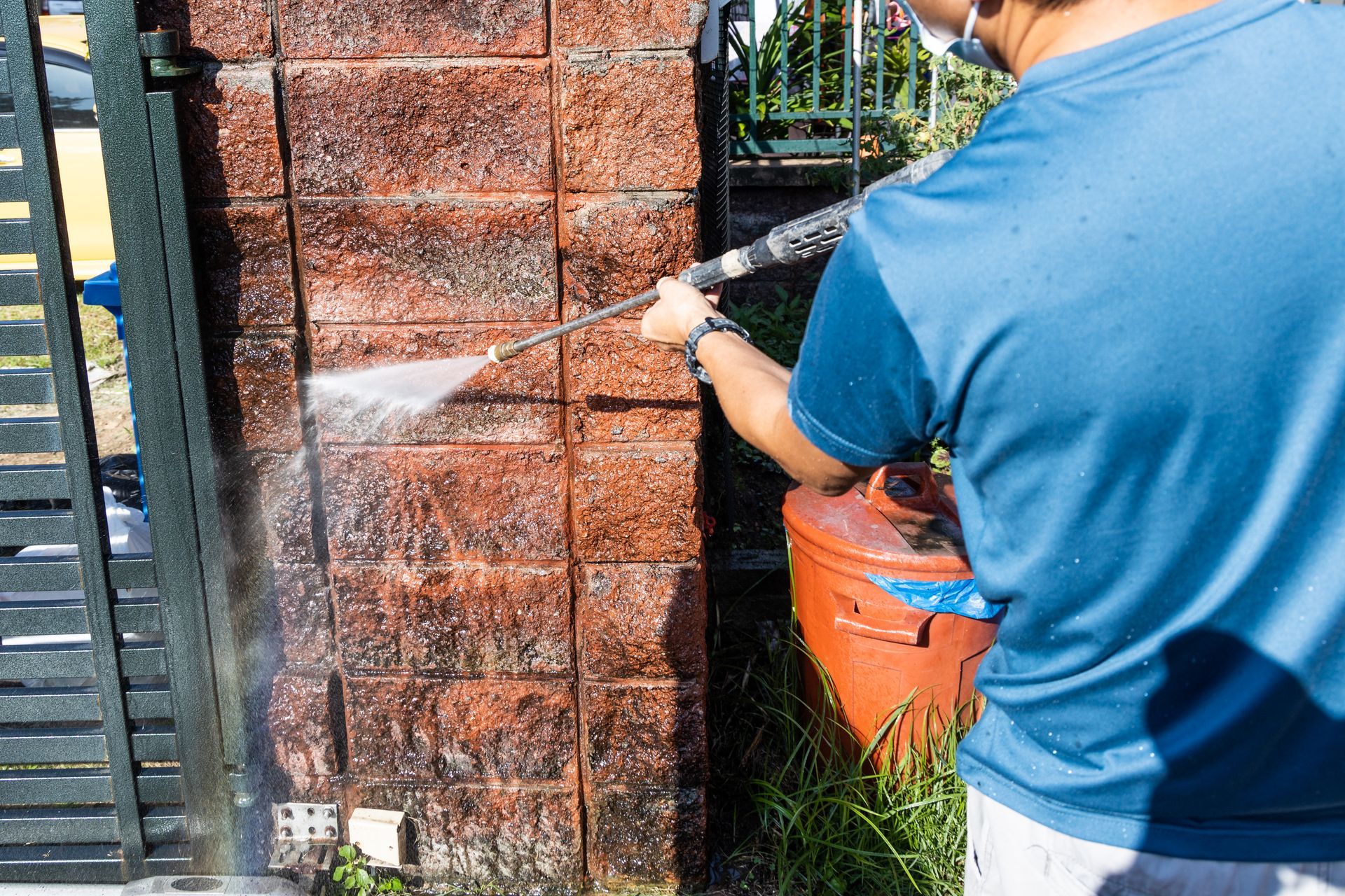 Person spray-washing a brick wall beside a gate with an orange pressure washer