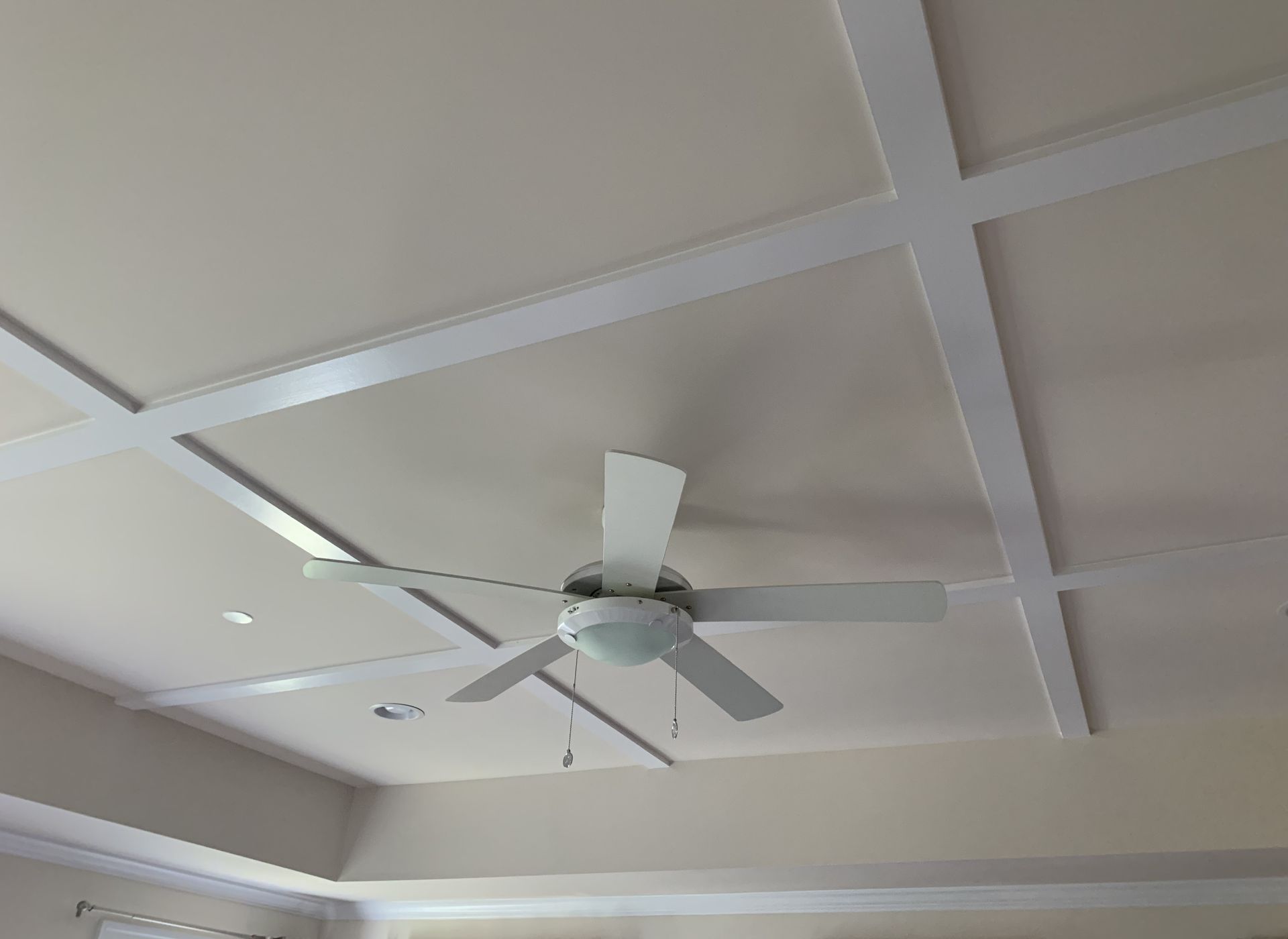White ceiling fan mounted on a coffered ceiling with white beams, in a room with light walls.