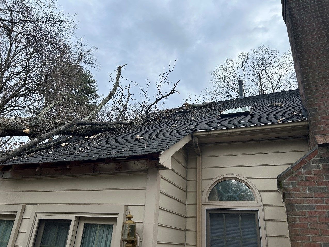 Tree branches lie across a house roof, damaging the shingles and skylight. The sky is overcast.