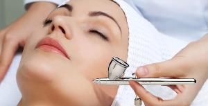 Woman receiving facial treatment with an airbrush; close-up of her face, eyes closed.